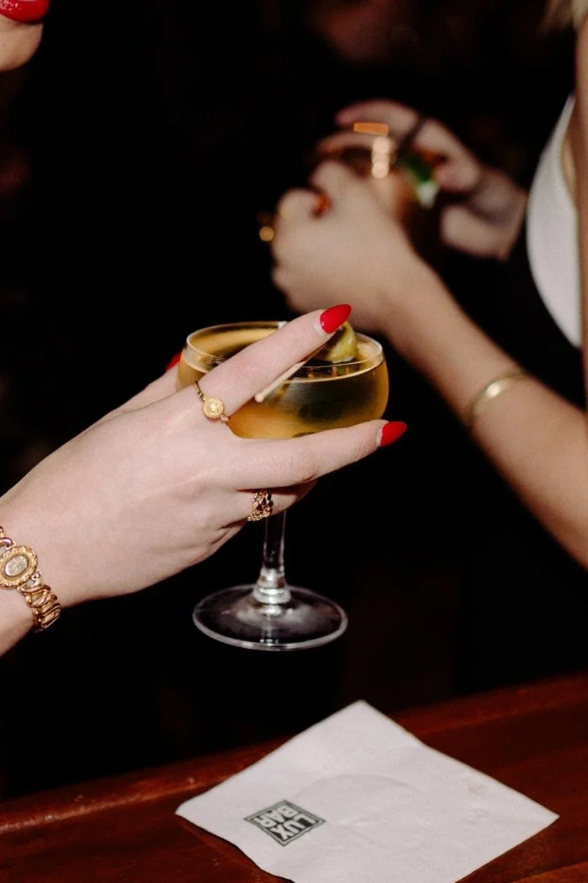 A hand with red-painted nails and gold jewelry holds a glass of white wine with a slice of lime inside at a bar or restaurant. Other people are visible in the background, and a paper napkin with a partially visible logo is on the wooden table.
