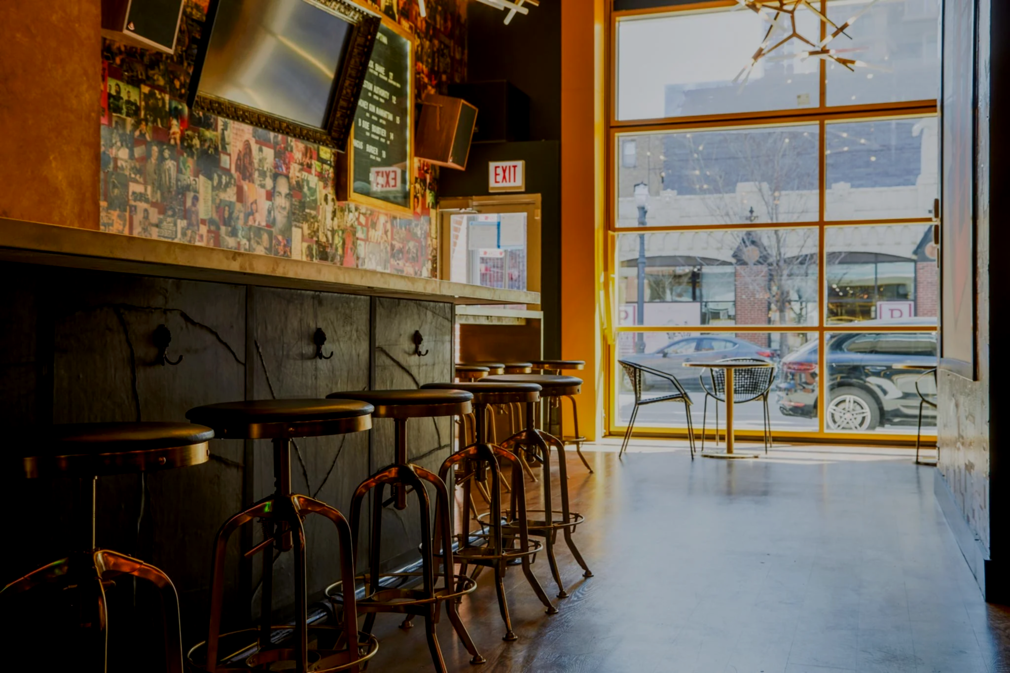 Interior of a cozy cafe with large windows, sunlight illuminating wooden chairs and a counter decorated with photos, and an exit door visible in the background.