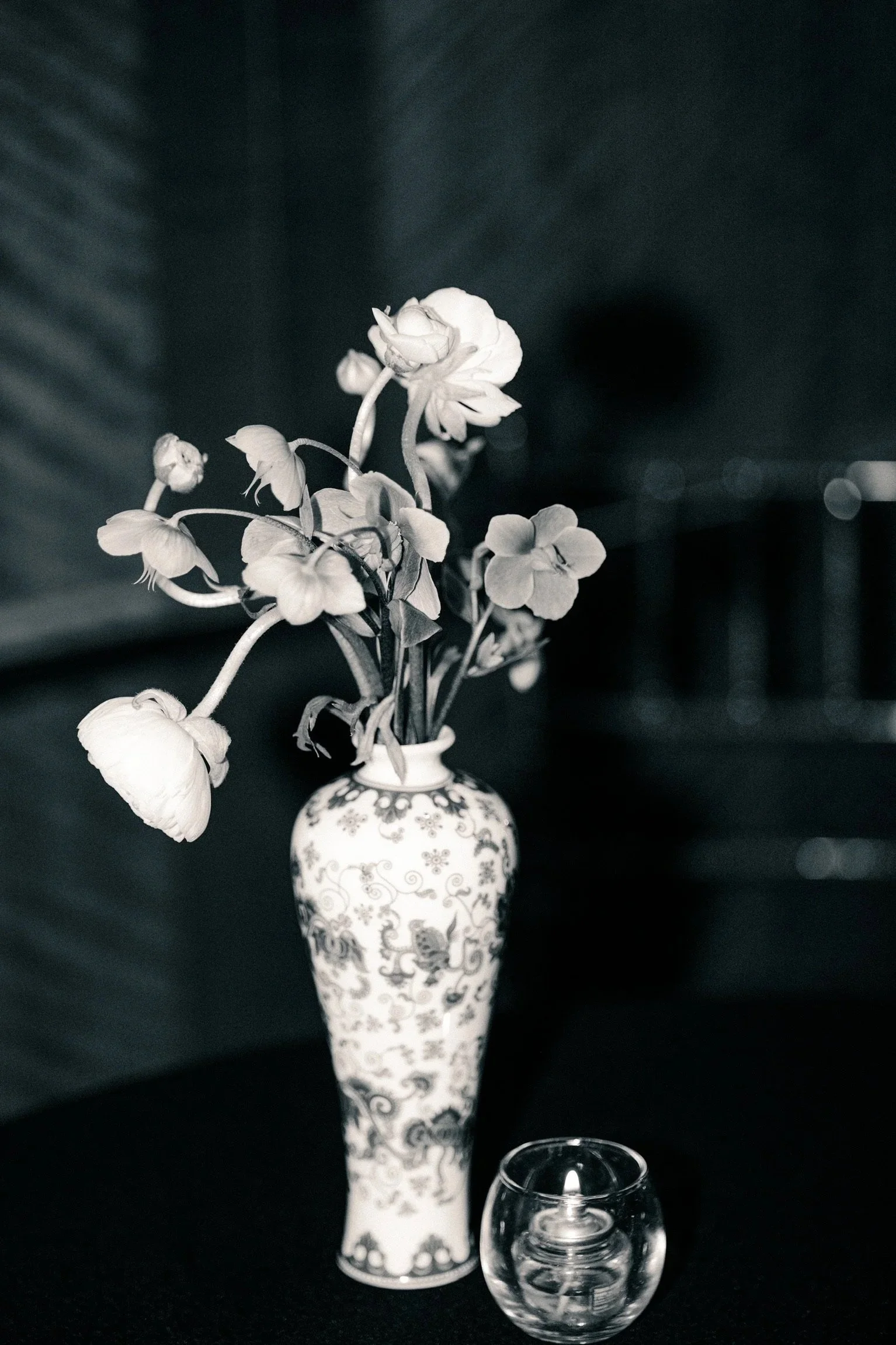 A decorative vase with a floral pattern holding a bouquet of flowers, with a glass of water in the foreground, placed on a dark surface against a dark background.