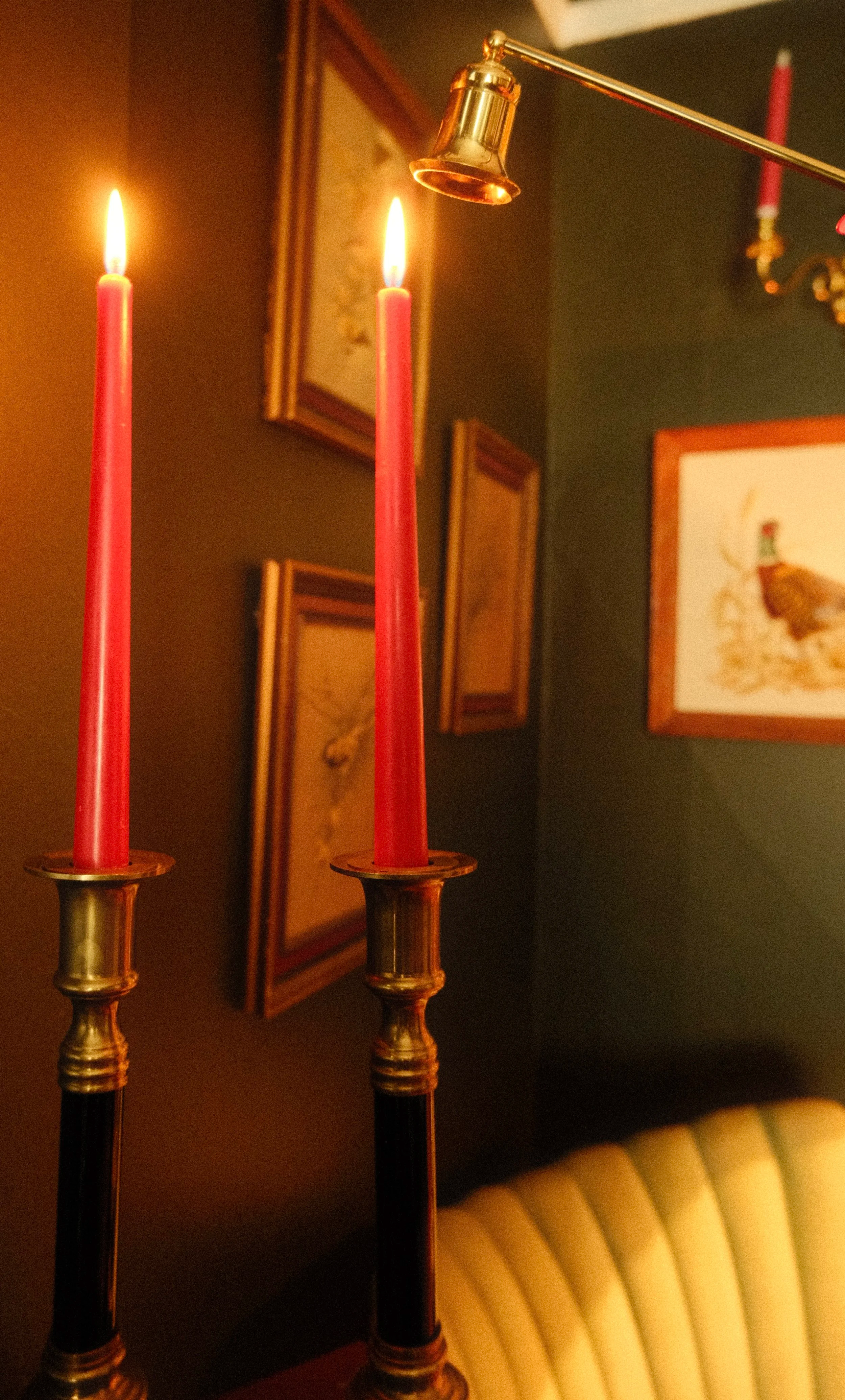 Two red candles burning in brass candle holders on a table with a dark-colored wall background. Framed artwork and a corner of a light-colored armchair are visible in the background.