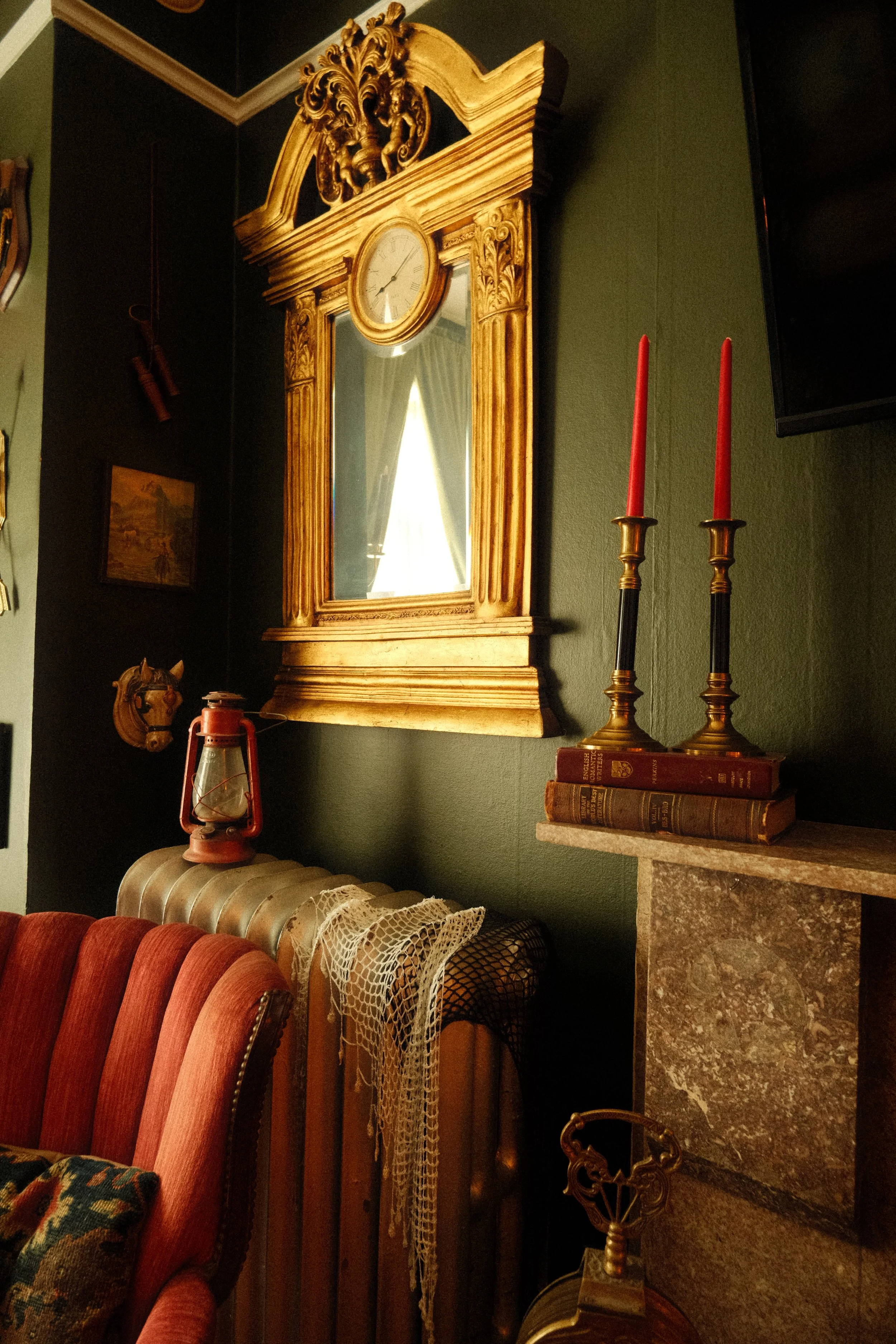Decorative gold-framed mirror with a clock at the top, two red candles in brass holders on a stack of vintage books, and antique items including a chest and a hunt cap in a cozy, dimly lit room.