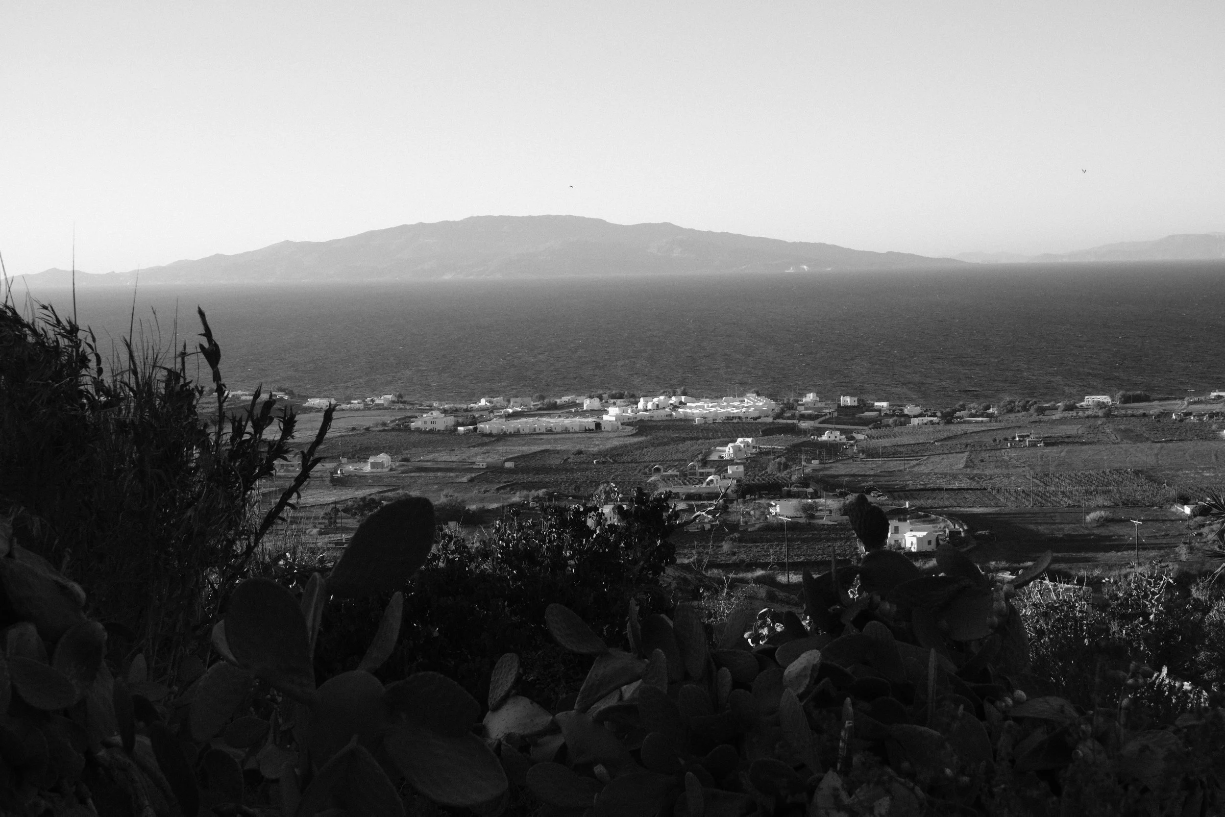 A black and white landscape showing a coastal town with buildings and fields in the foreground, a large body of water behind it, and an island or mountain in the distance under a clear sky.