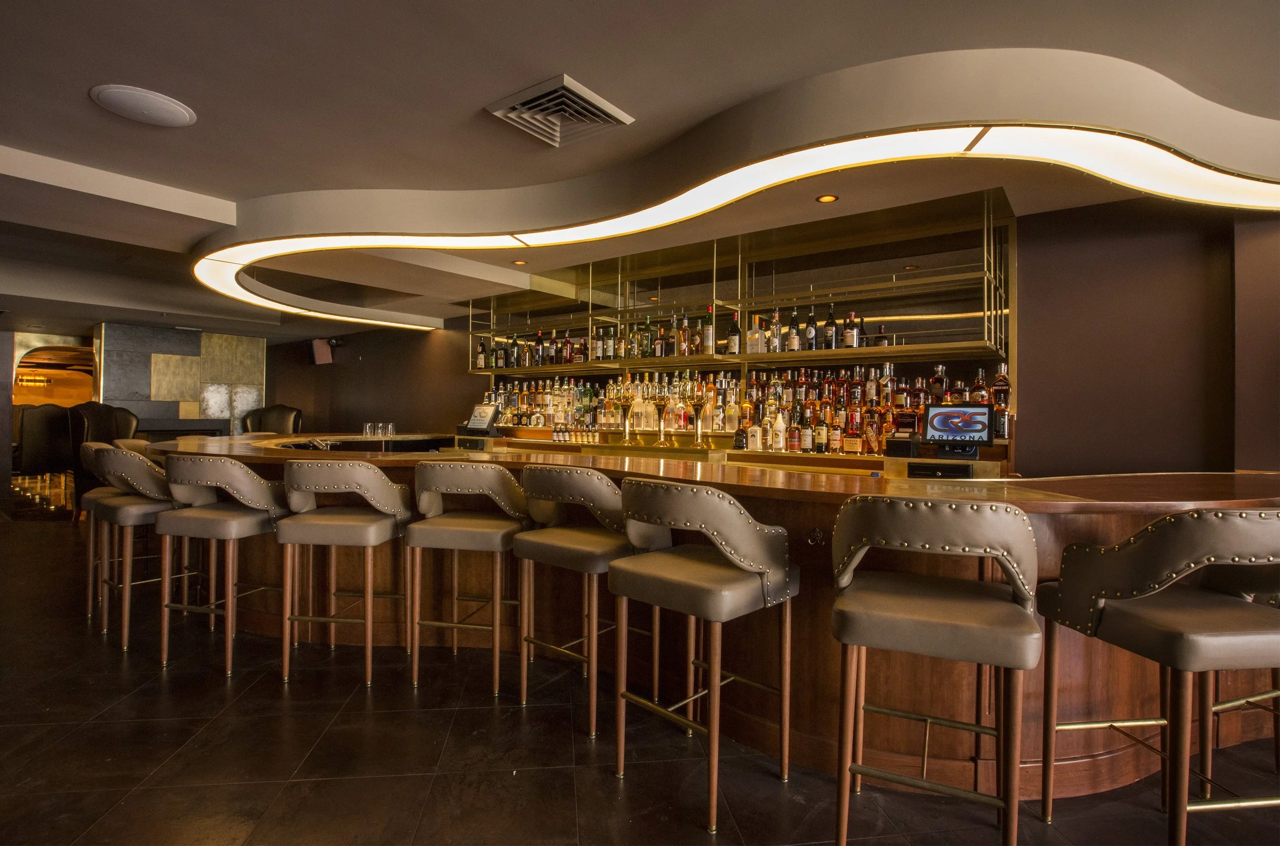 A modern bar with a curved ceiling light fixture, a bar counter with various liquor bottles, and cushioned bar stools.