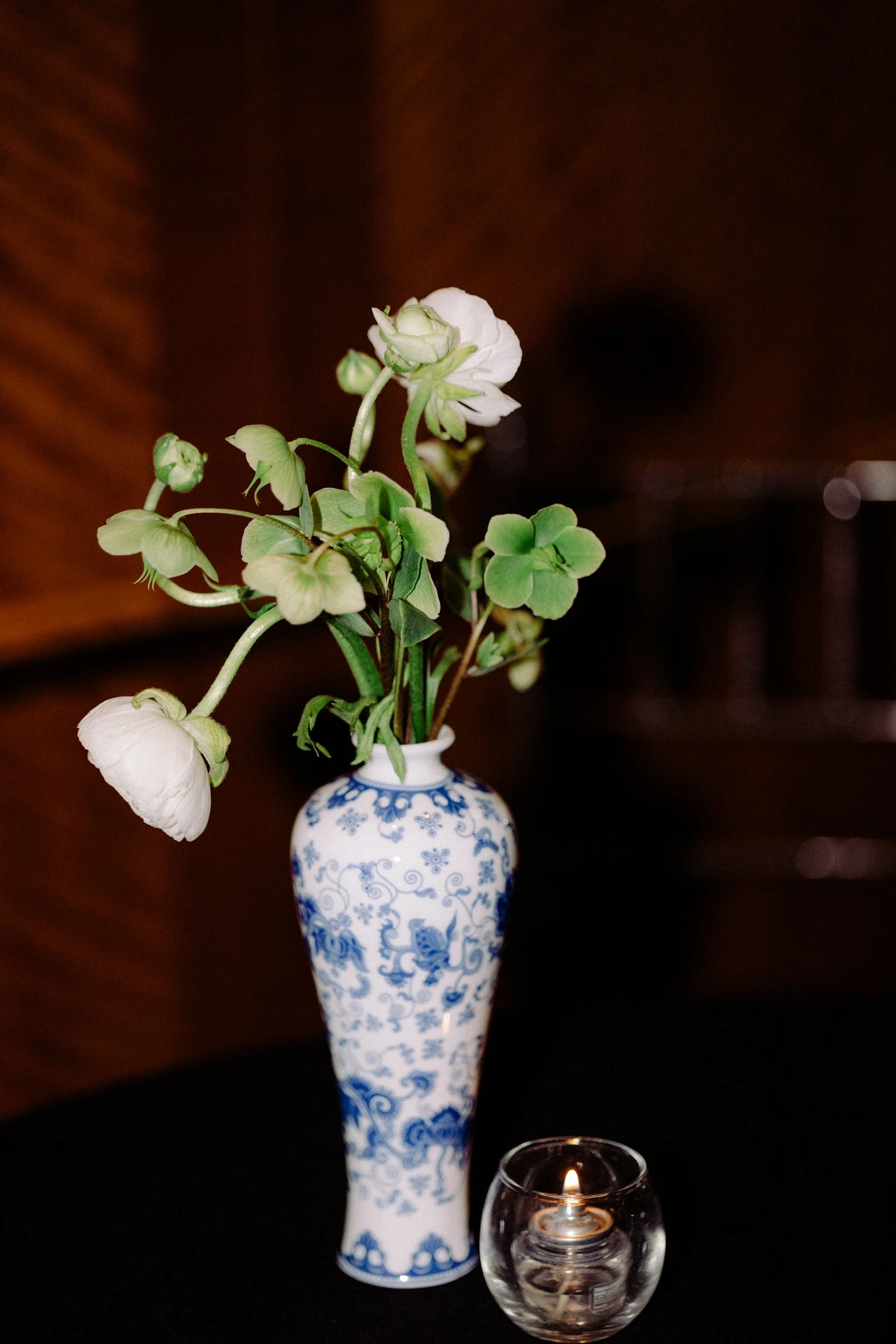 A decorative white and blue porcelain vase with floral patterns, holding white flowering plants. A small glass votive candle holder with a lit candle is placed in front of the vase on a dark surface.