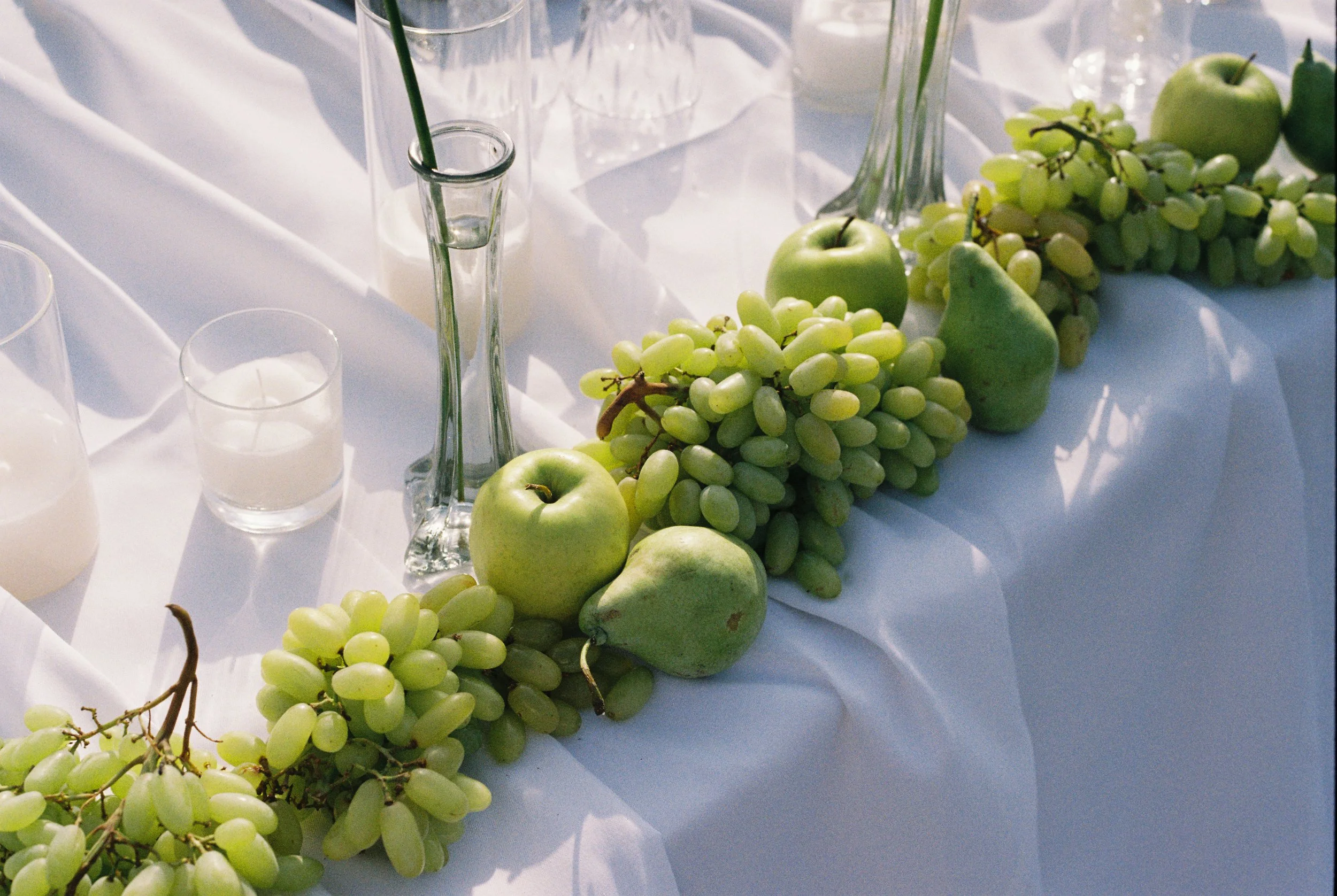 A table decorated with green apples, green pears, bunches of green grapes, and several glass candle holders with white candles on a white tablecloth.