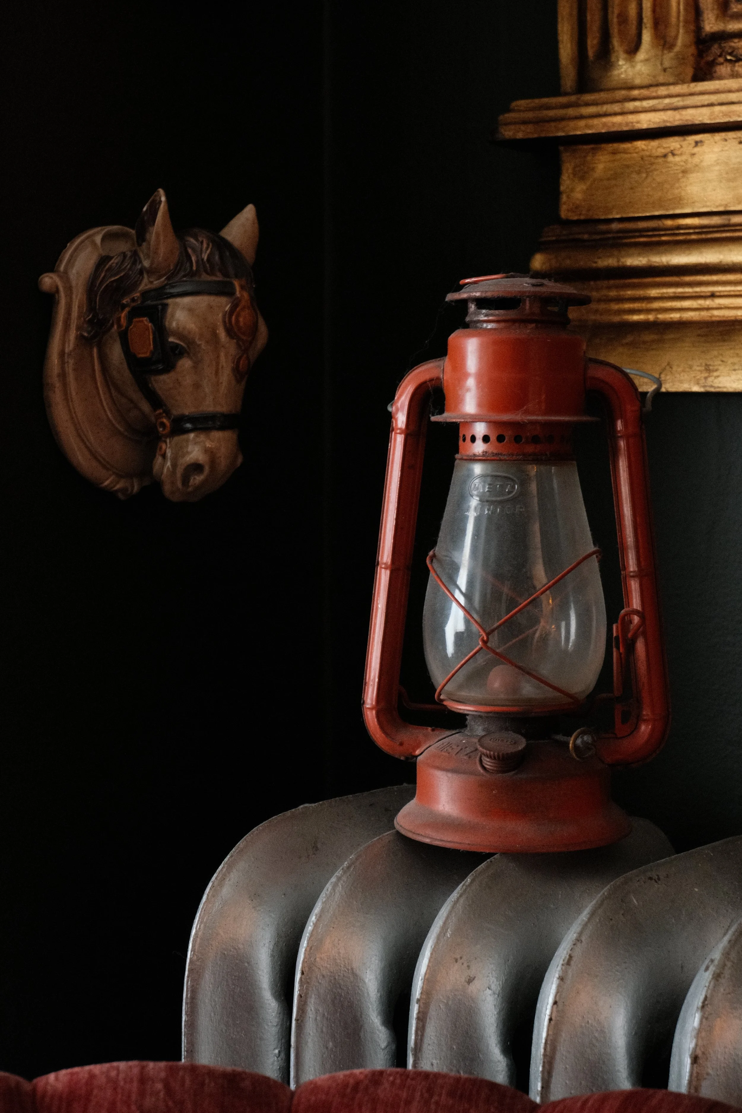 A wooden horse head wall decoration with a black eyepiece, an aged red lantern, and three vintage metal plates stacked together, all set against a dark background.