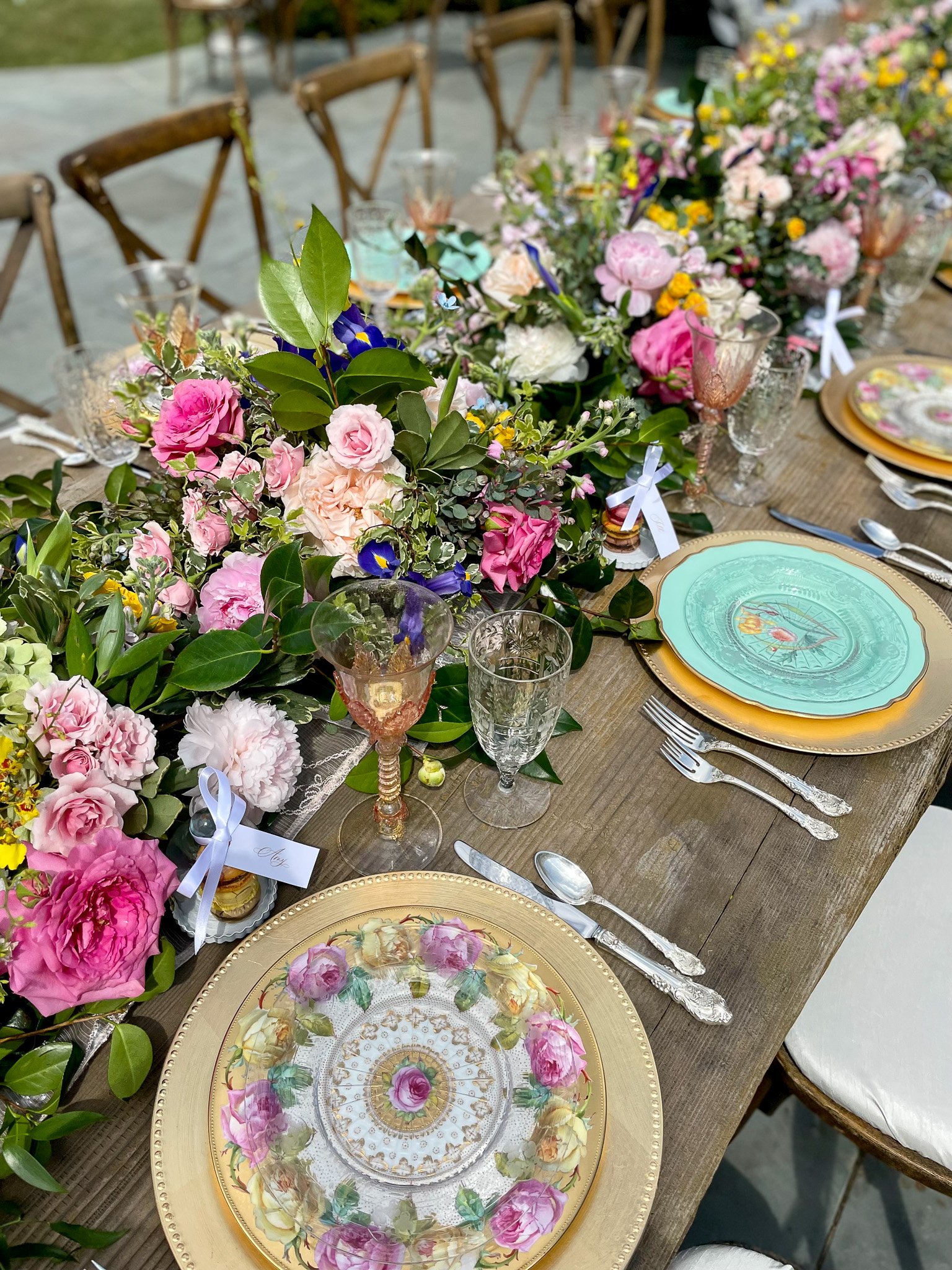 Elegant outdoor dining table decorated with floral arrangements, fine china with floral patterns, crystal glasses, and silverware, set for a celebration or special occasion.