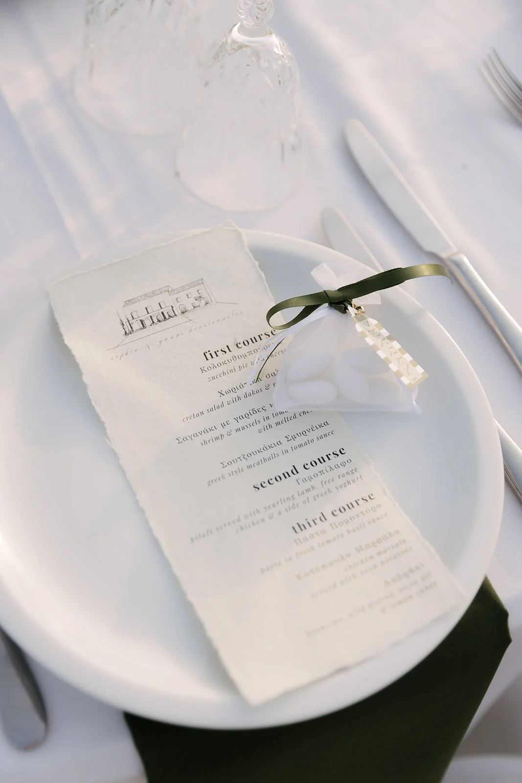 A white plate holding a wedding menu card, a small favor bag decorated with a ribbon, a knife and fork, and two wine glasses on a table with a white tablecloth.