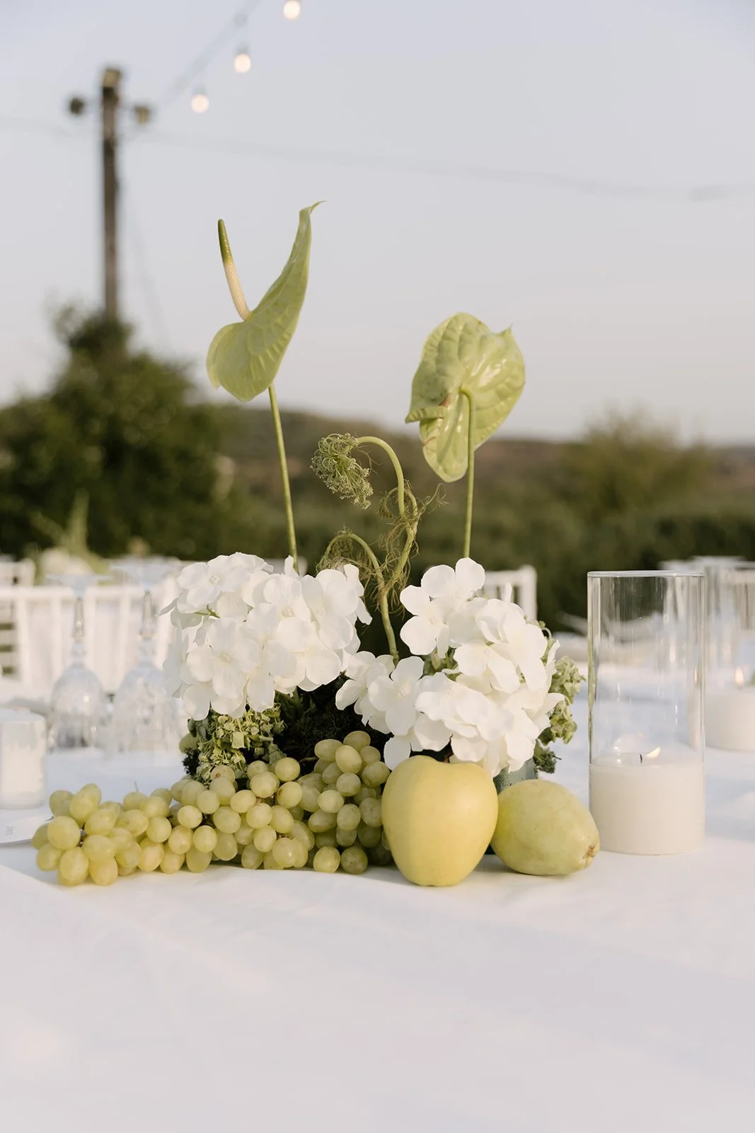 A floral centerpiece with white hydrangeas, green apples, and grapes, with decorative green anthurium flowers and candles, on a white table at an outdoor event.