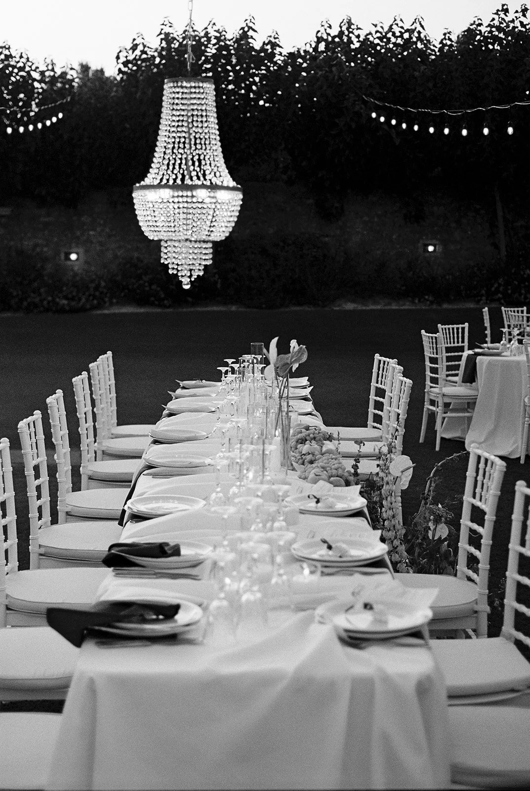 Outdoor dining setup for an event with elegantly arranged table, chairs, and a large chandelier hanging overhead, surrounded by string lights at dusk.