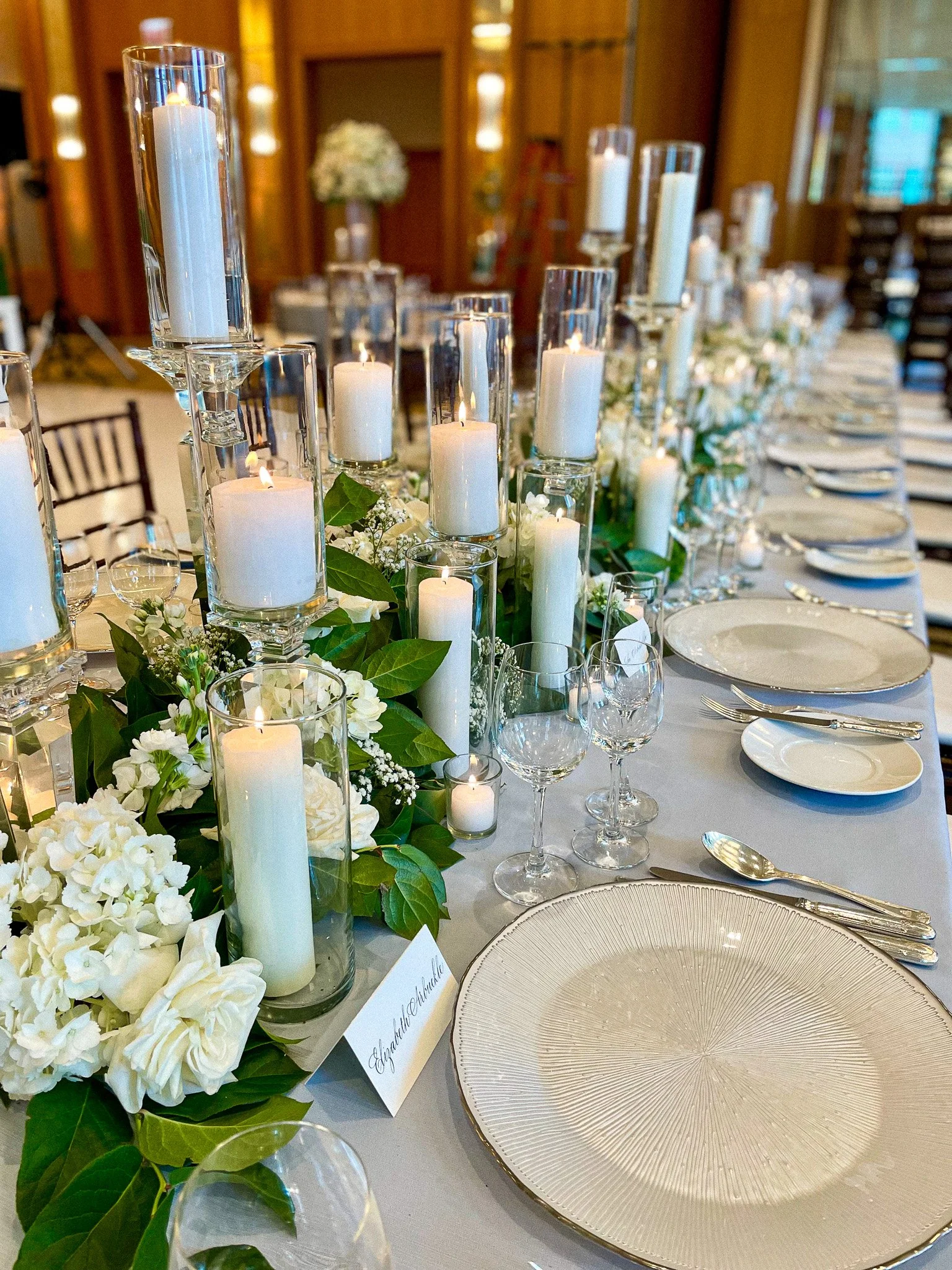 Elegant banquet table decorated with white candles in glass holders and greenery, set with white plates and glassware, in a richly decorated room with wooden walls.