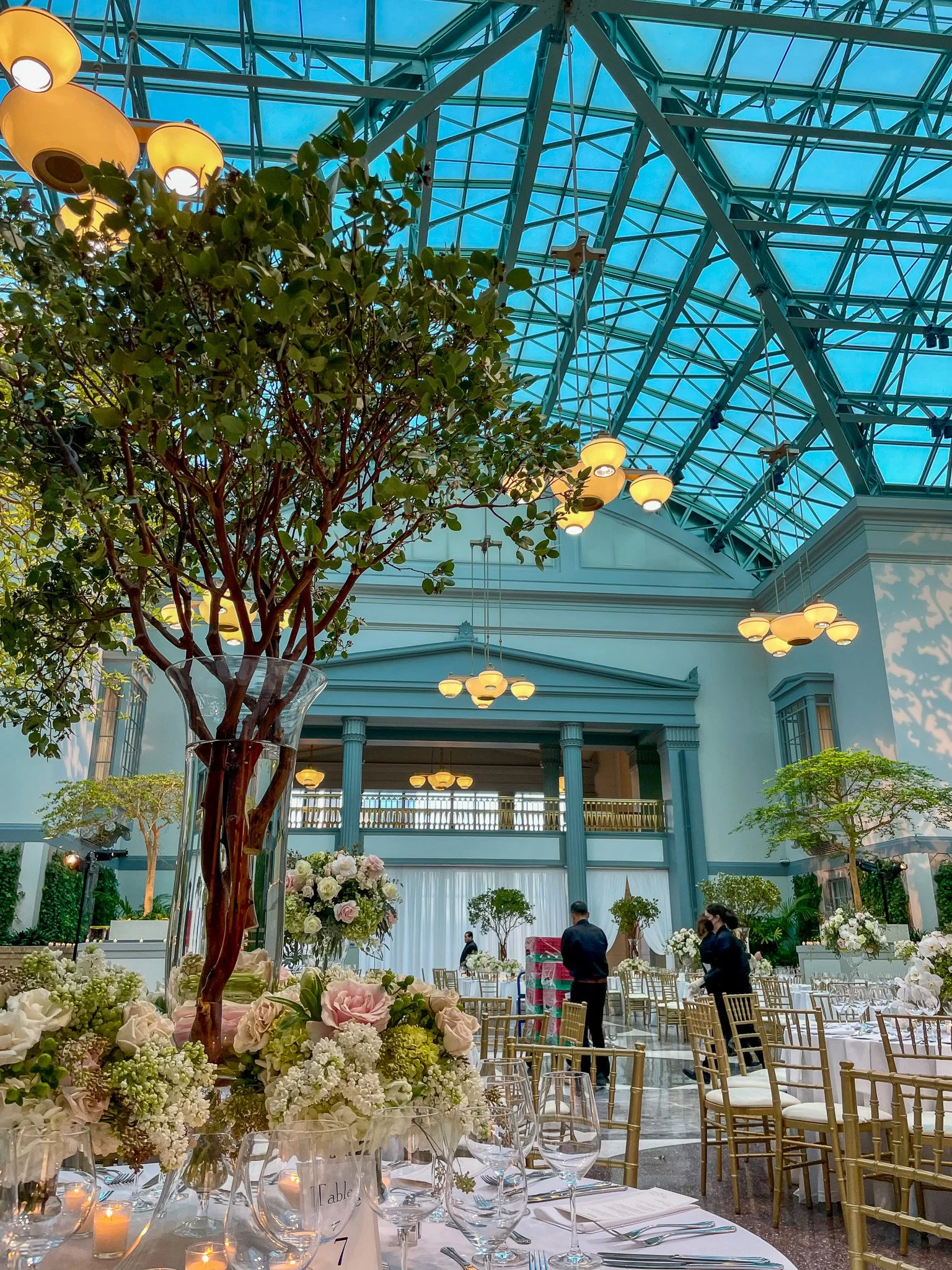 An elegant event or wedding reception hall decorated with floral centerpieces, high ceiling with glass panels, green trees, and warm lighting, with a few people preparing tables.