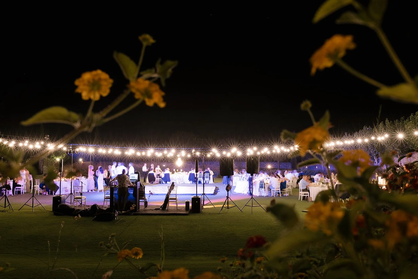 Outdoor nighttime wedding reception under string lights with guests seated at decorated tables and a dance floor with a DJ setup in the foreground.