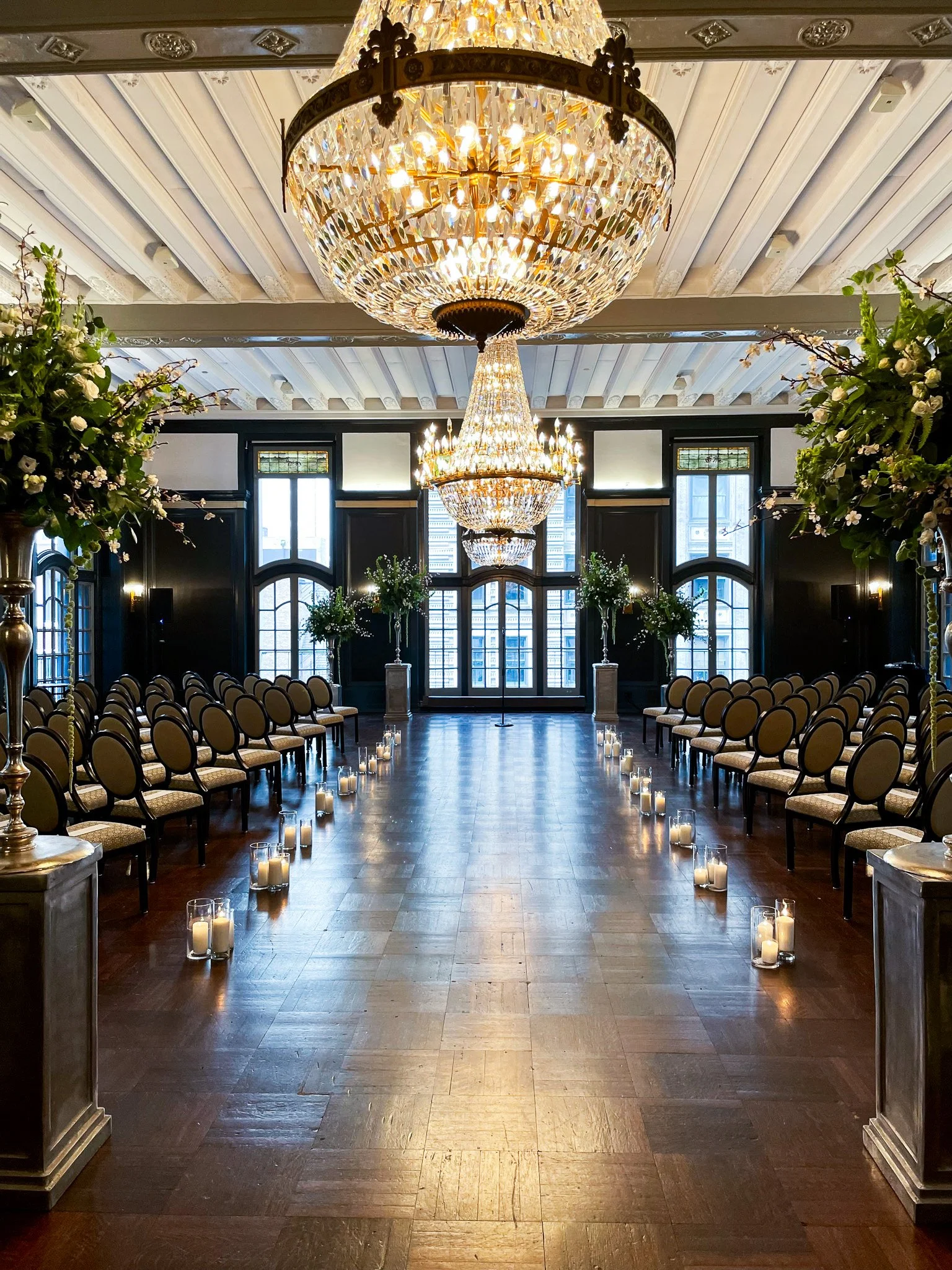Elegant wedding ceremony setup inside a grand hall with a high ceiling, multiple large chandeliers, rows of chairs, flower arrangements, and candles lining the aisle.