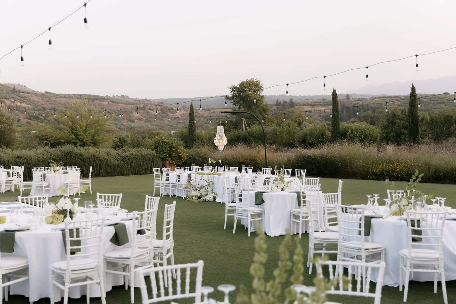 Outdoor wedding reception setup with round tables draped in white tablecloths, white chairs, and floral centerpieces, set on a green lawn with string lights overhead, surrounded by trees and hills.