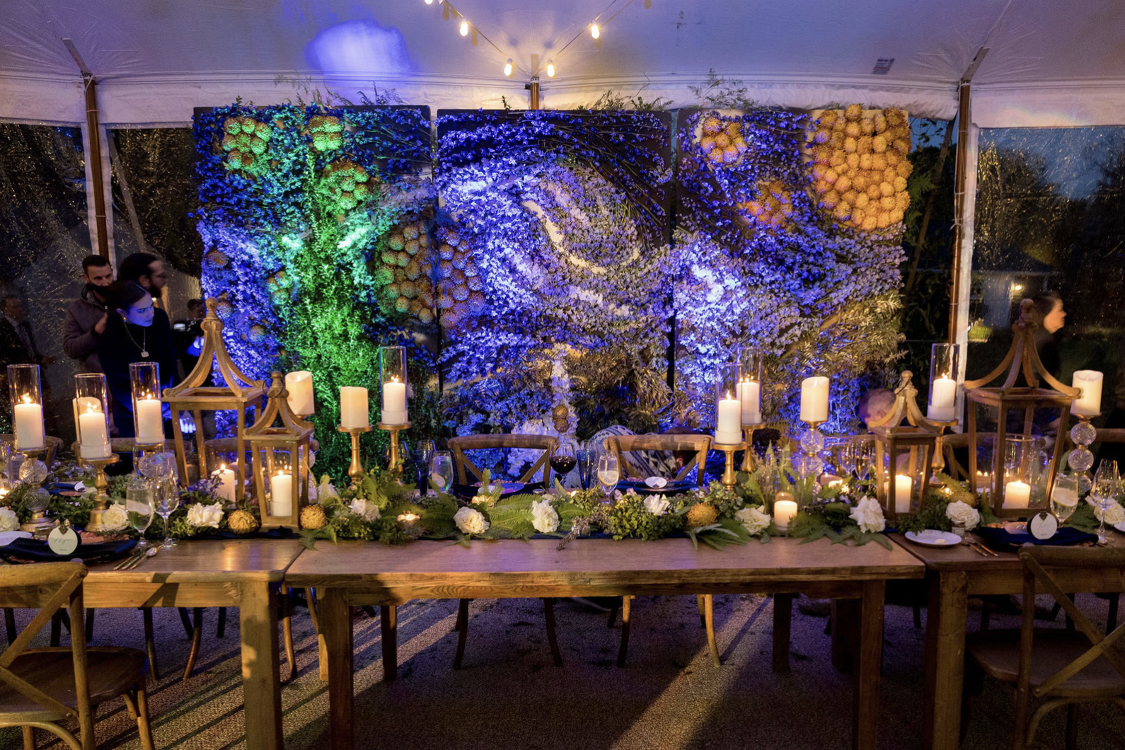Decorated event table with candles, flowers, and lanterns inside a tent with a floral wall and string lights.