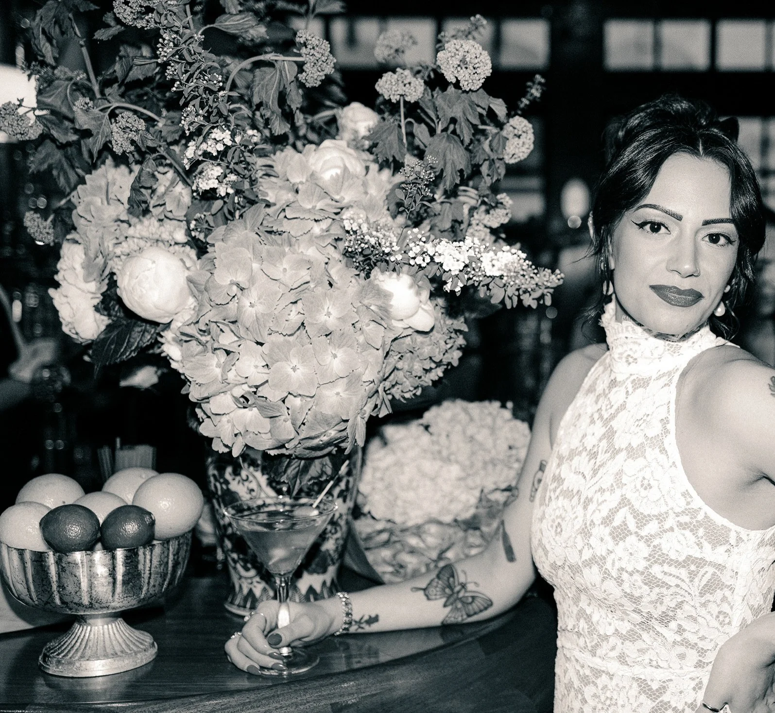 A woman with dark hair styled in loose waves, wearing a lace top, standing at a bar or table with a large bouquet of flowers and a bowl of fruit.