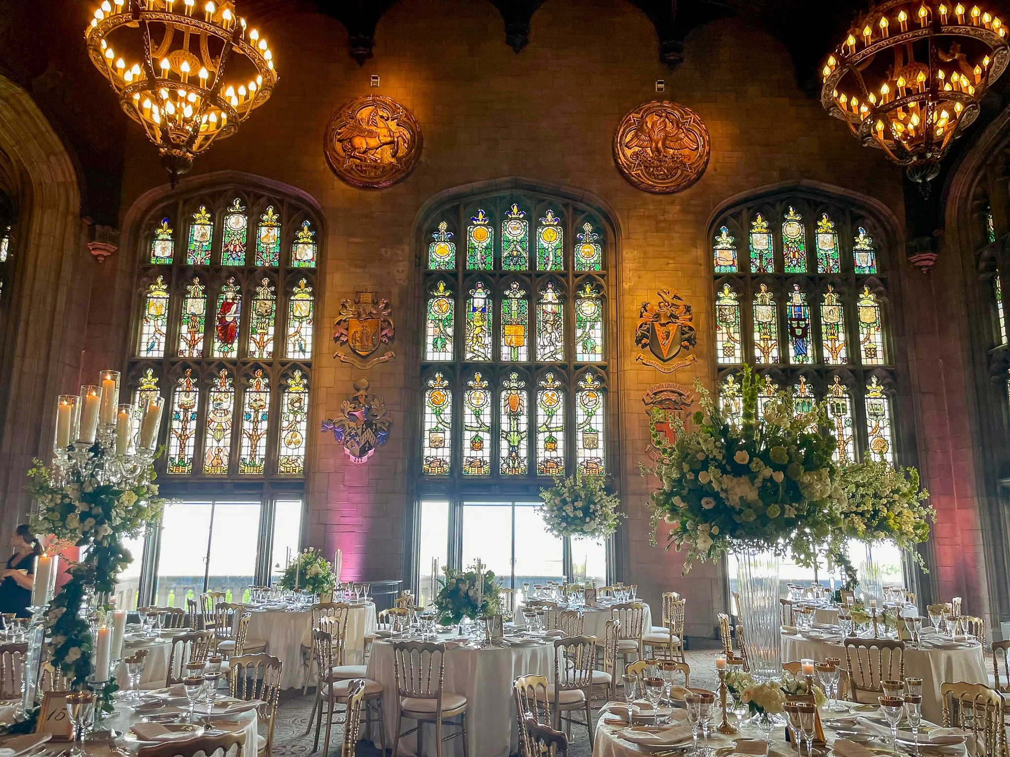 Interior of a grand hall decorated for a formal event, with round tables covered in white tablecloths, elegant chair arrangements, large floral centerpieces, many lit candles, and stained glass windows on the back wall. There are chandeliers hanging 
