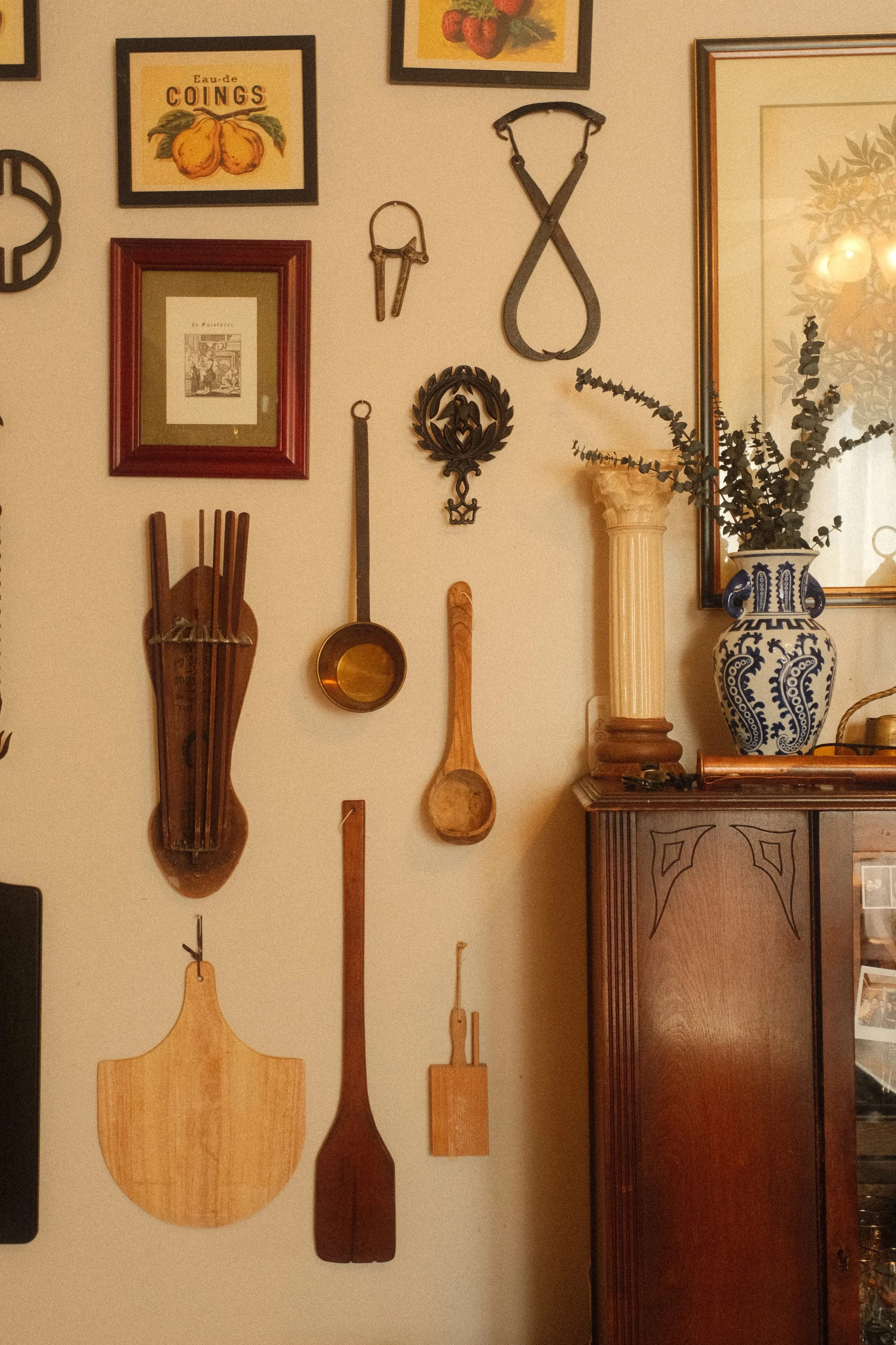 Wall decorated with framed artwork, antique courtship tools, and wooden paddles, including a large ceramic vase with greenery and a small wooden table.