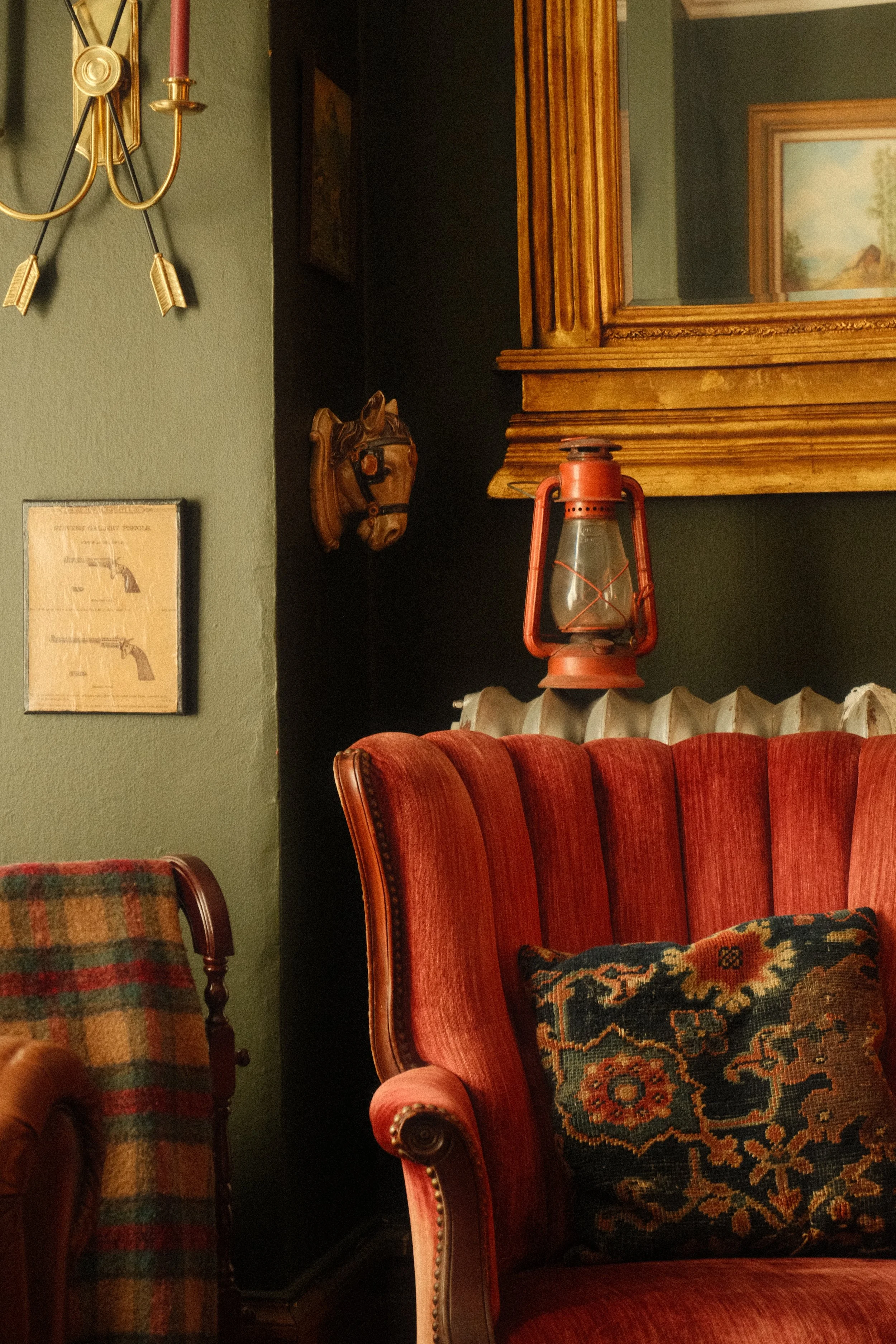 A vintage living room corner with a high back red upholstered armchair, a wooden chair with a plaid blanket, a framed illustration of guns, a horse head wall sculpture, a wooden cabinet with glass, a red oil lantern, a radiator, and a framed landscap