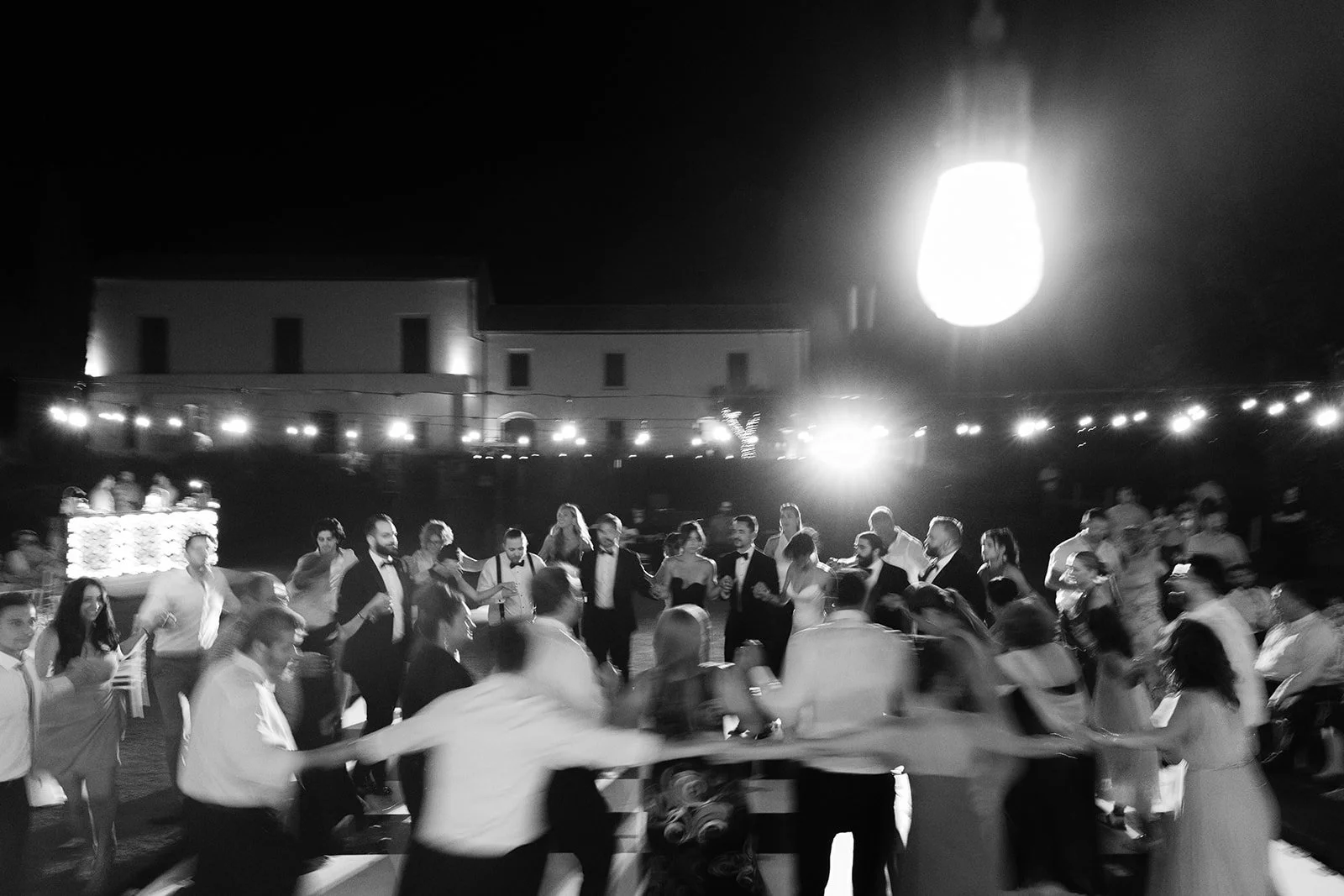 People dancing and socializing at a nighttime outdoor wedding reception, with string lights and a large bright light in the background.