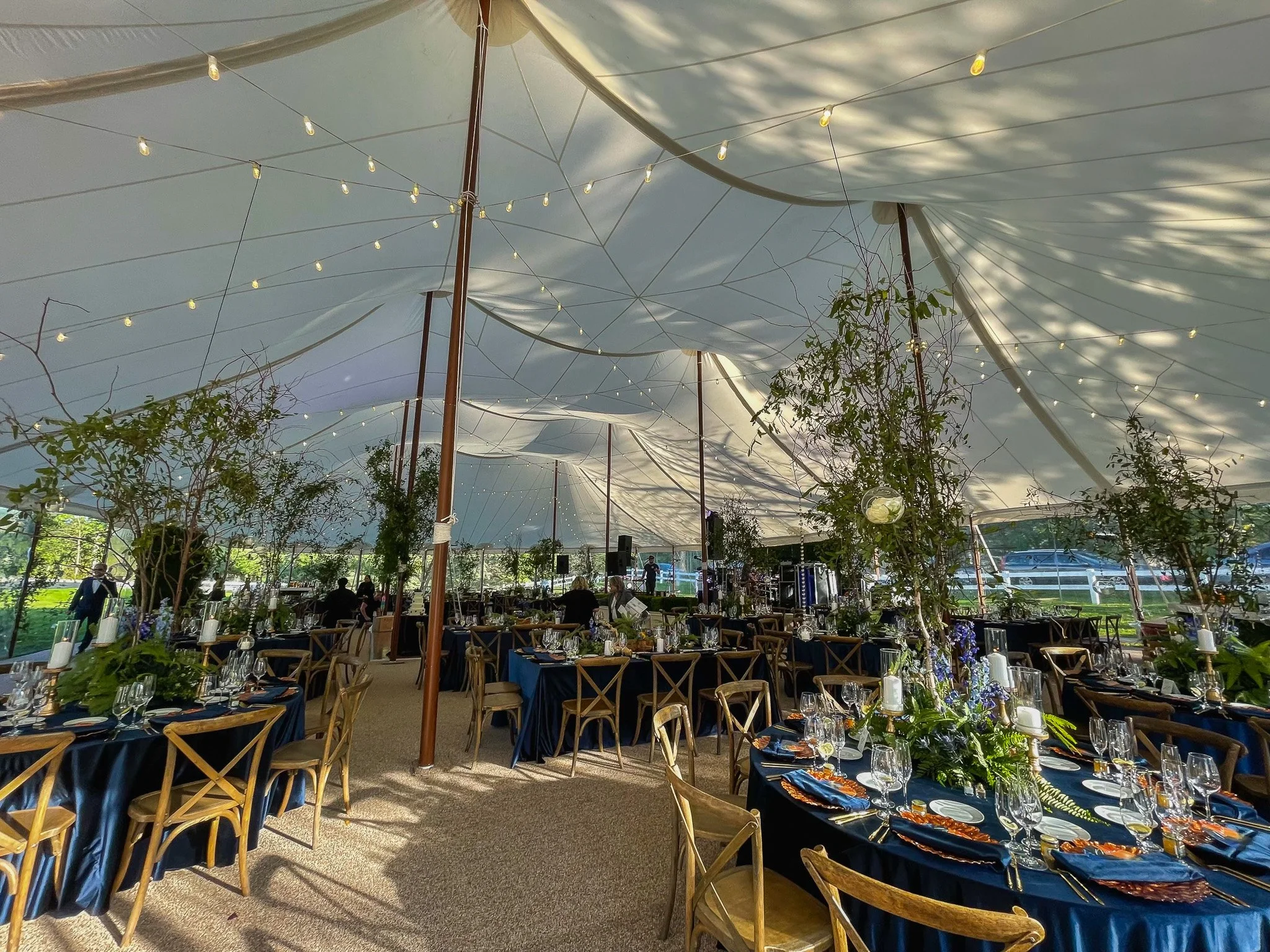 Indoor event space with tables set for a formal dinner, decorated with navy tablecloths, glassware, and floral centerpieces. The ceiling is covered with large white fabric drapes and string lights, creating a festive atmosphere. Outside light filters