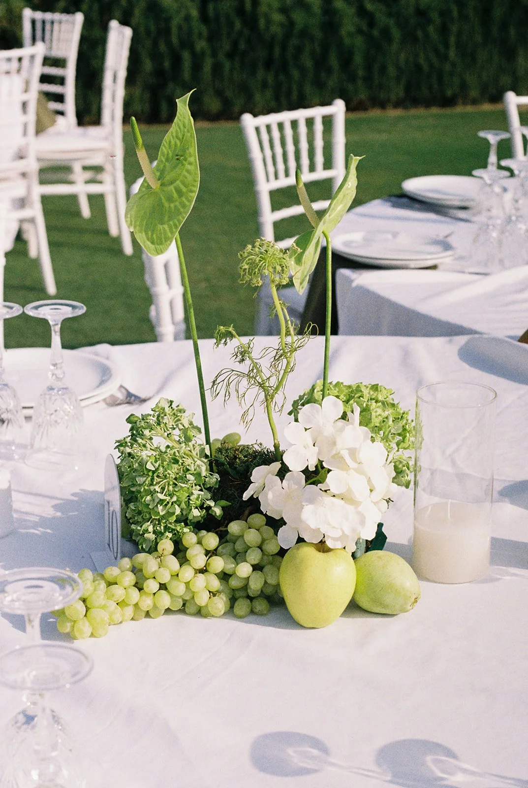 Elegant outdoor table centerpiece with green apples, white hydrangeas, and tropical leaves, set on a white tablecloth with white chairs.