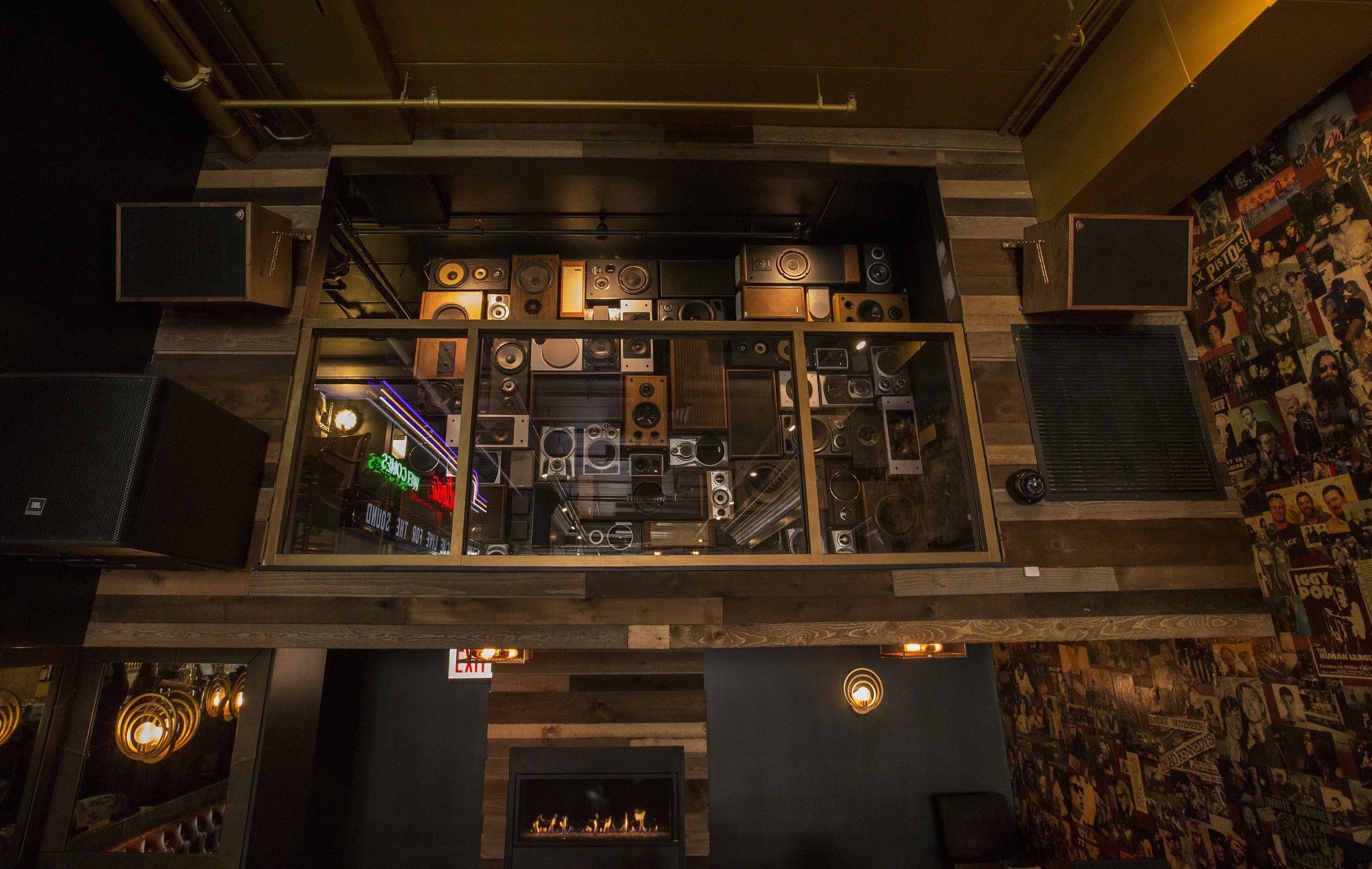 Interior of a room with a collection of vintage speakers on the wall and a framed glass display case below. The room has wood-paneled walls and a mural with various figures and music posters on the right side.