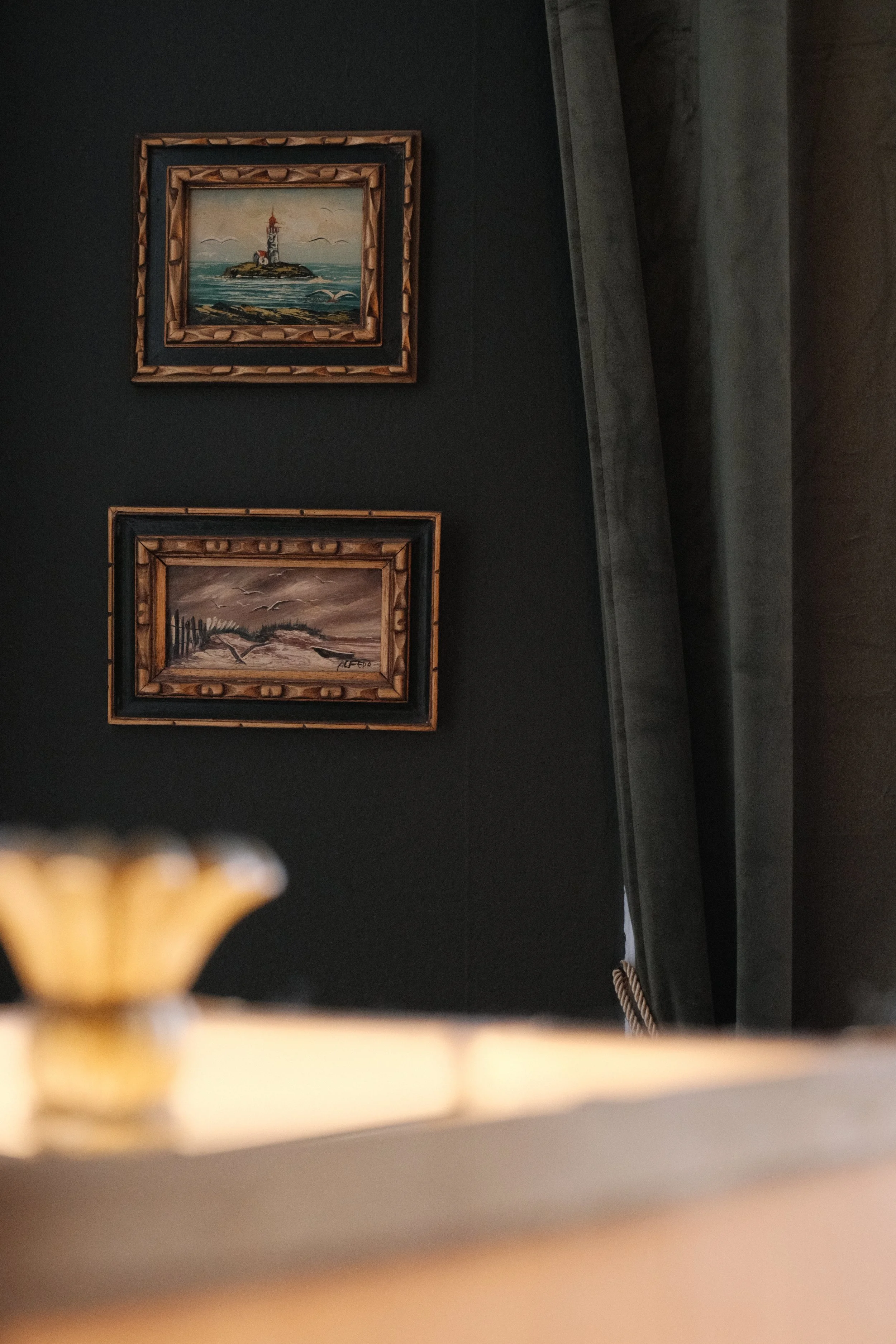Two framed paintings hanging on a dark-colored wall, one above the other, with a curtain on the right side of the image and part of a wooden object visible at the bottom.