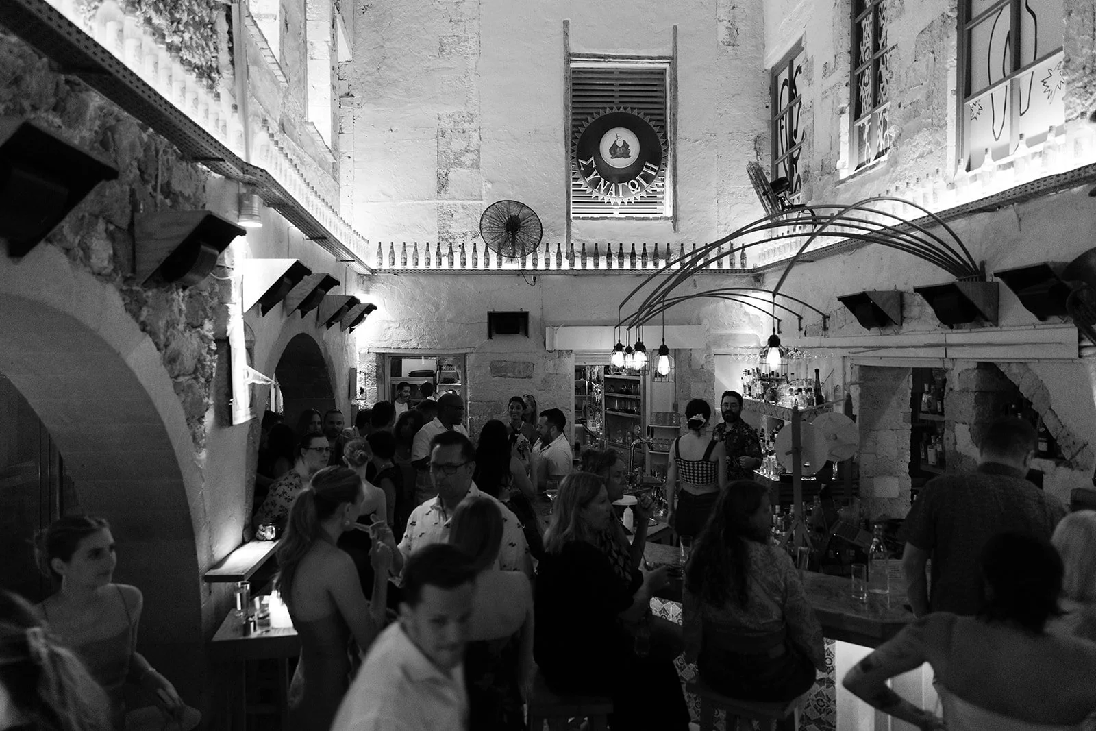 People socializing inside a rustic restaurant or bar with exposed brick walls, a balcony, and dim lighting.