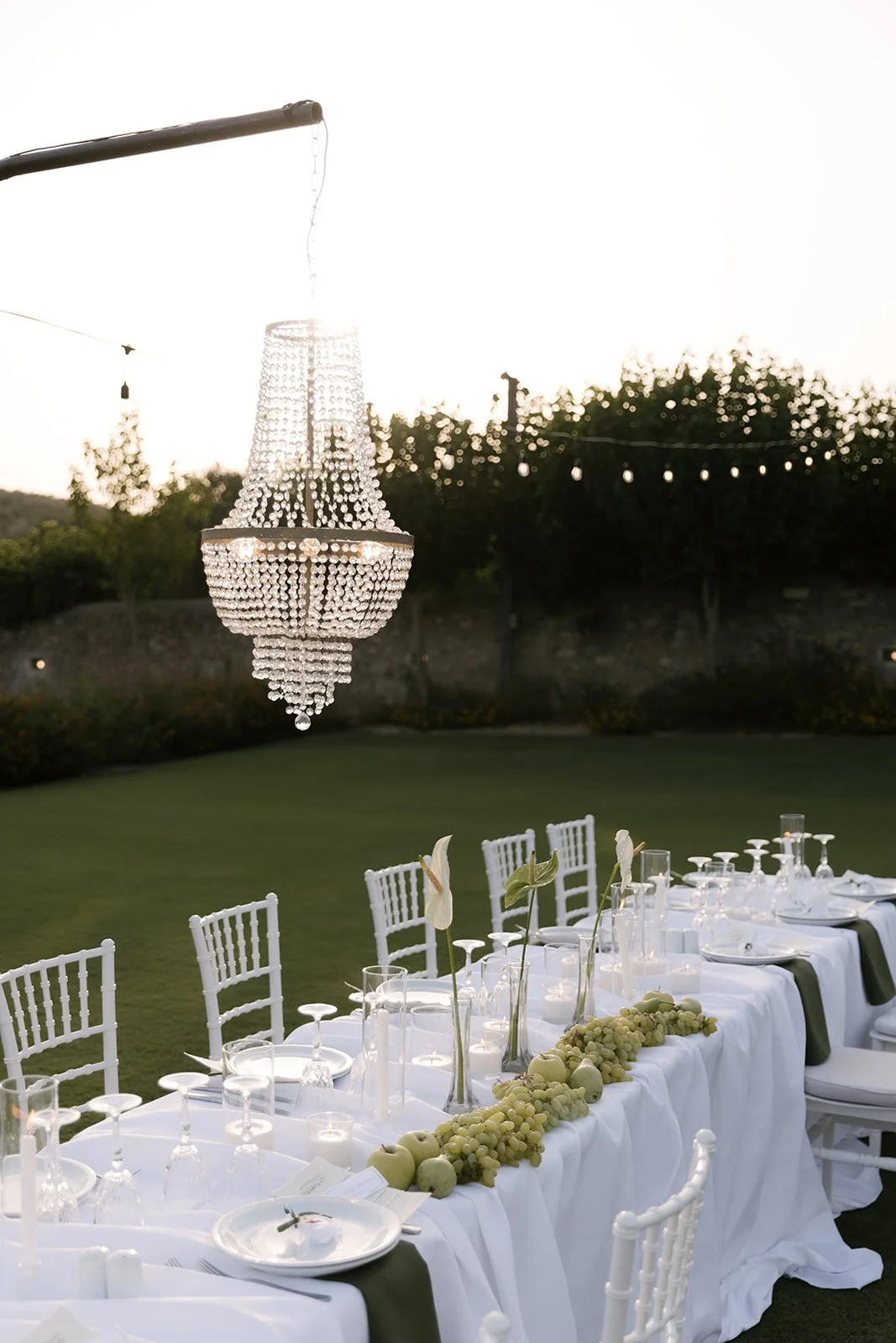 Outdoor dinner table decorated with white tablecloth, green napkins, white plates, wine glasses, and candles, set under a chandelier with hanging crystal beads, with white chairs and a row of grapes and apples as centerpieces, in a garden at sunset.