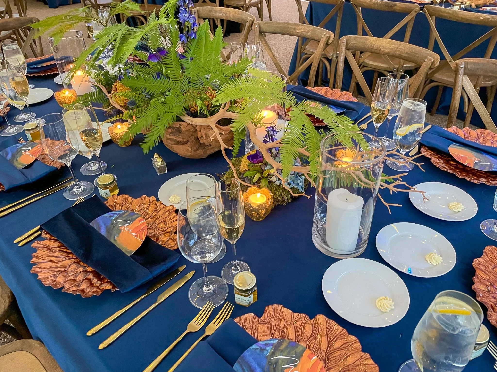A banquet table decorated with a large central floral arrangement of green ferns and purple flowers, surrounded by lit candles, champagne glasses, and orange and navy napkins on a dark blue tablecloth.