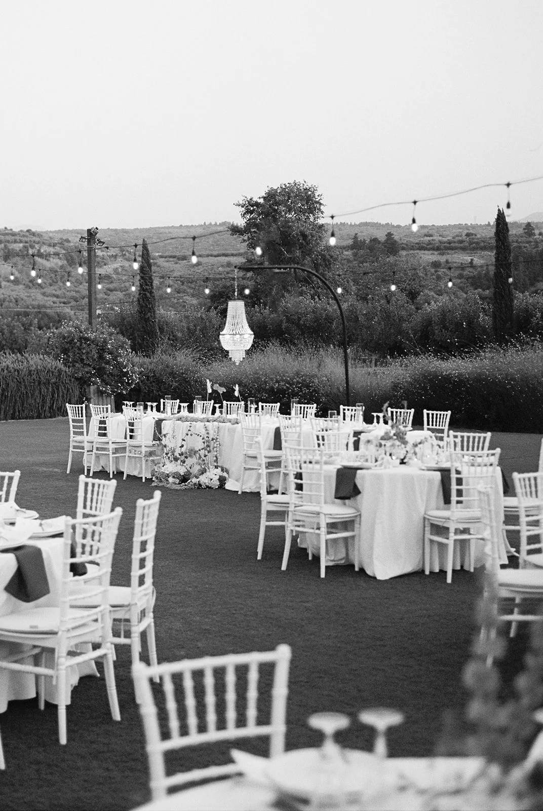 Outdoor banquet setup with round tables and white chairs, floral centerpieces, string lights overhead, and a chandelier hanging in the background, with a natural landscape and trees.