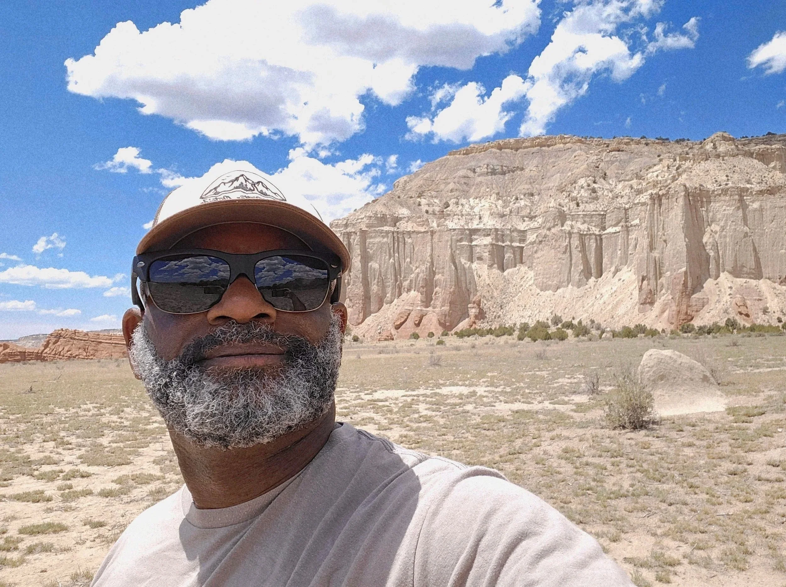 A man with glasses and a baseball cap taking a selfie outdoors near cliffs with a partly cloudy sky.