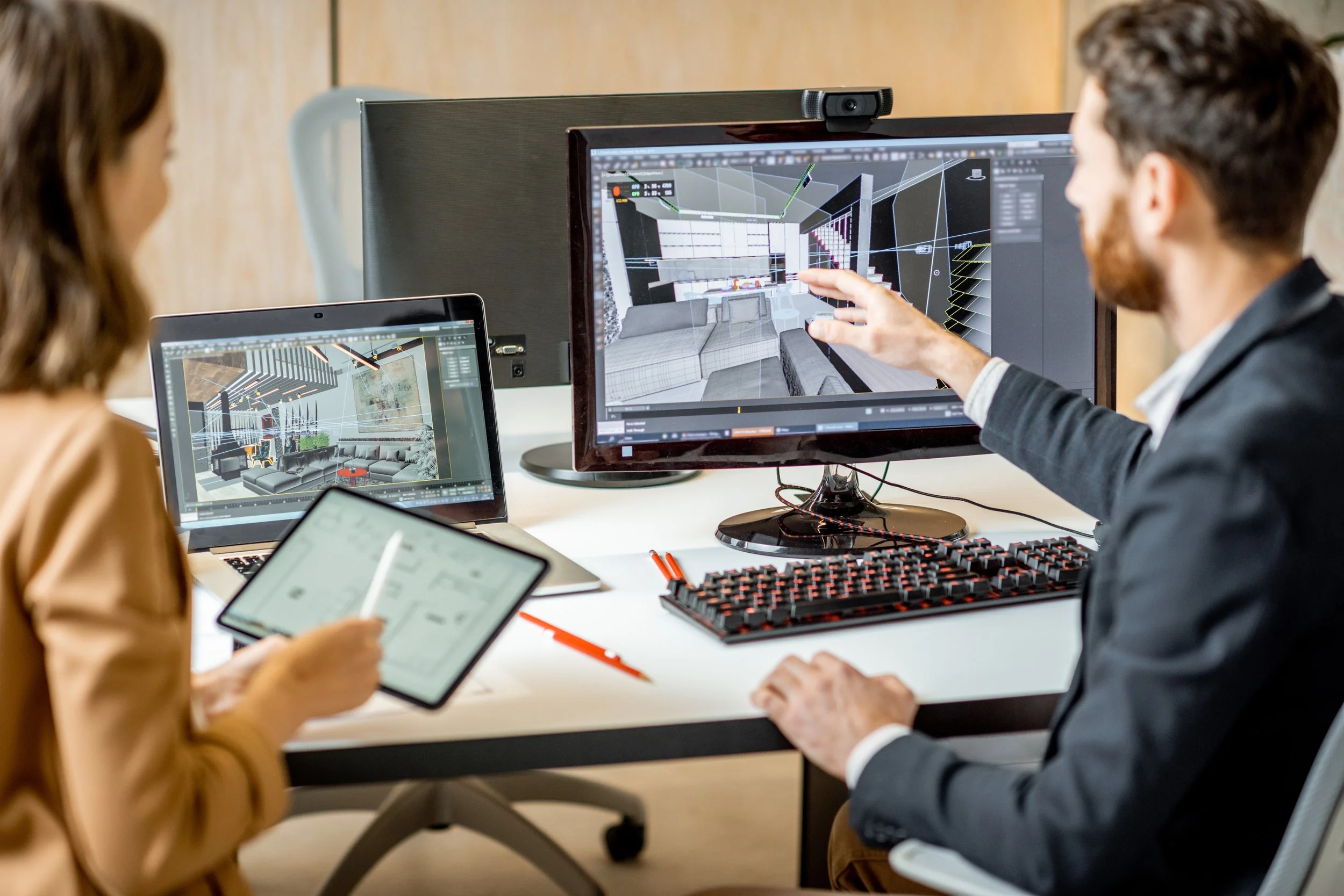 Two people working on interior design projects at a desk, with a computer and tablet showing 3D layouts of a living room and interior space.