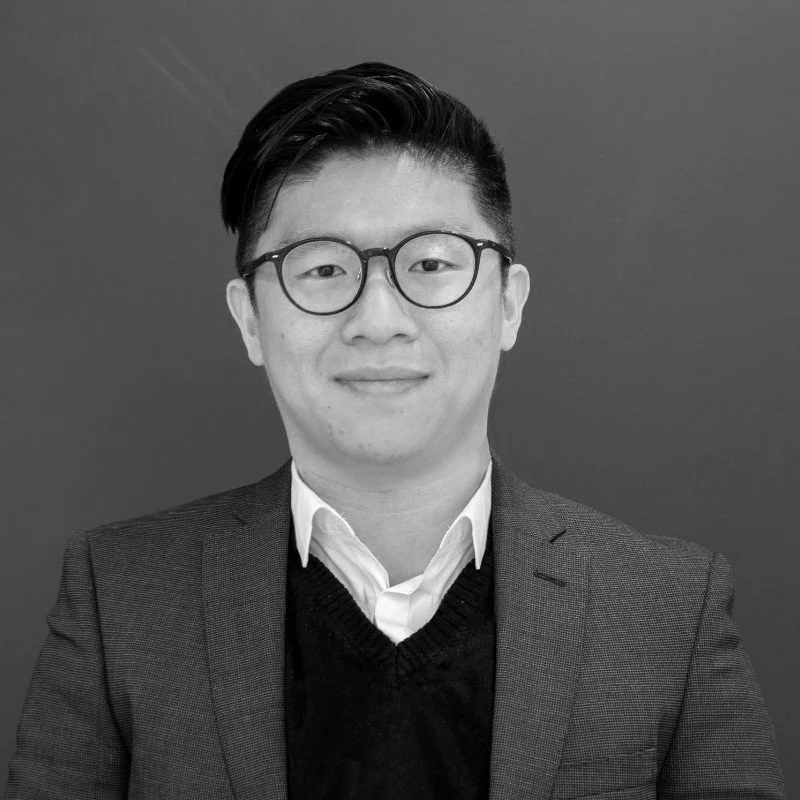 Black and white portrait of a young architect wearing glasses, a white collared shirt, and a dark sweater under a blazer, standing against a plain background.