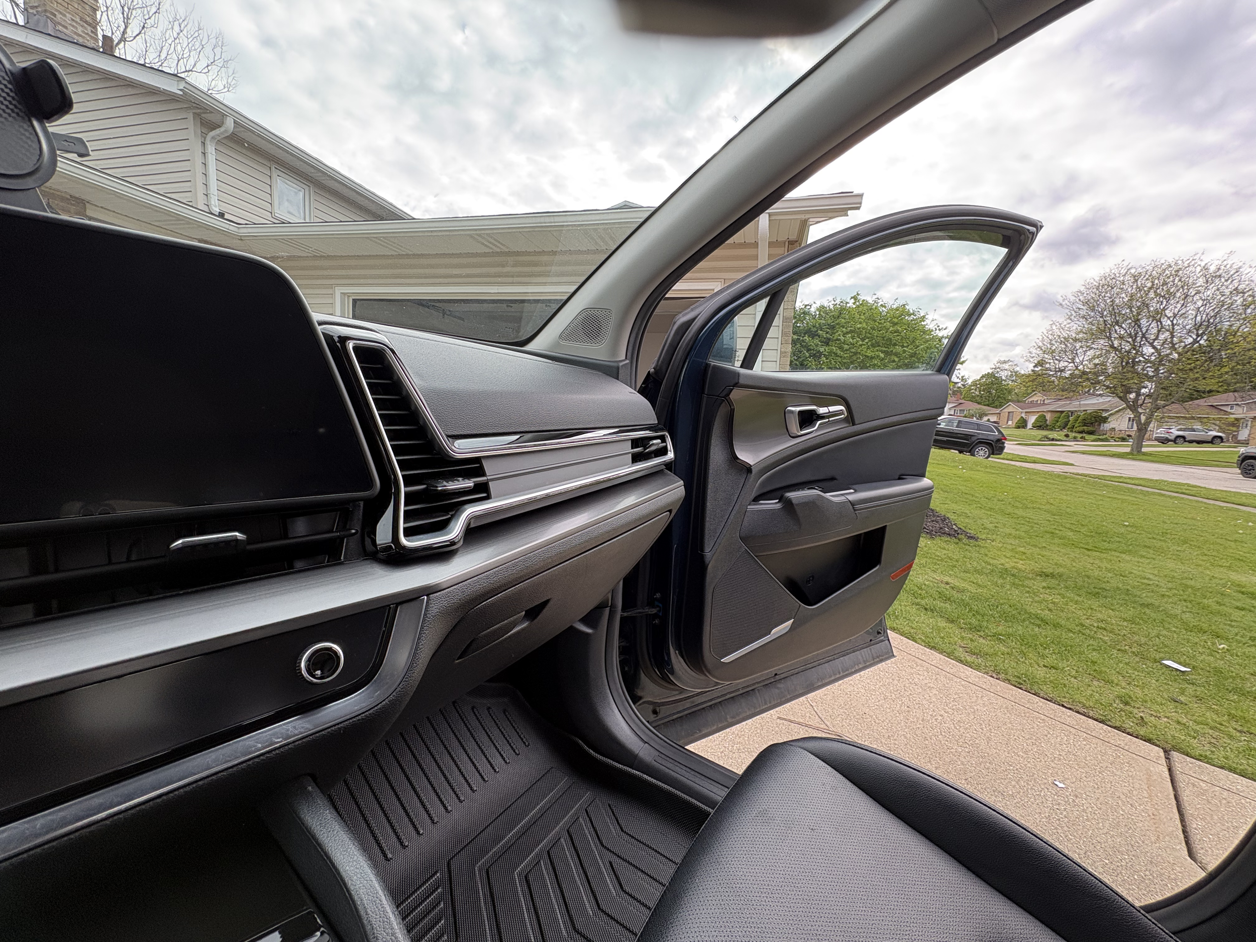 Inside a vehicle, showing the dashboard and the open front passenger door, with a view of residential houses, trees, and a cloudy sky outside.