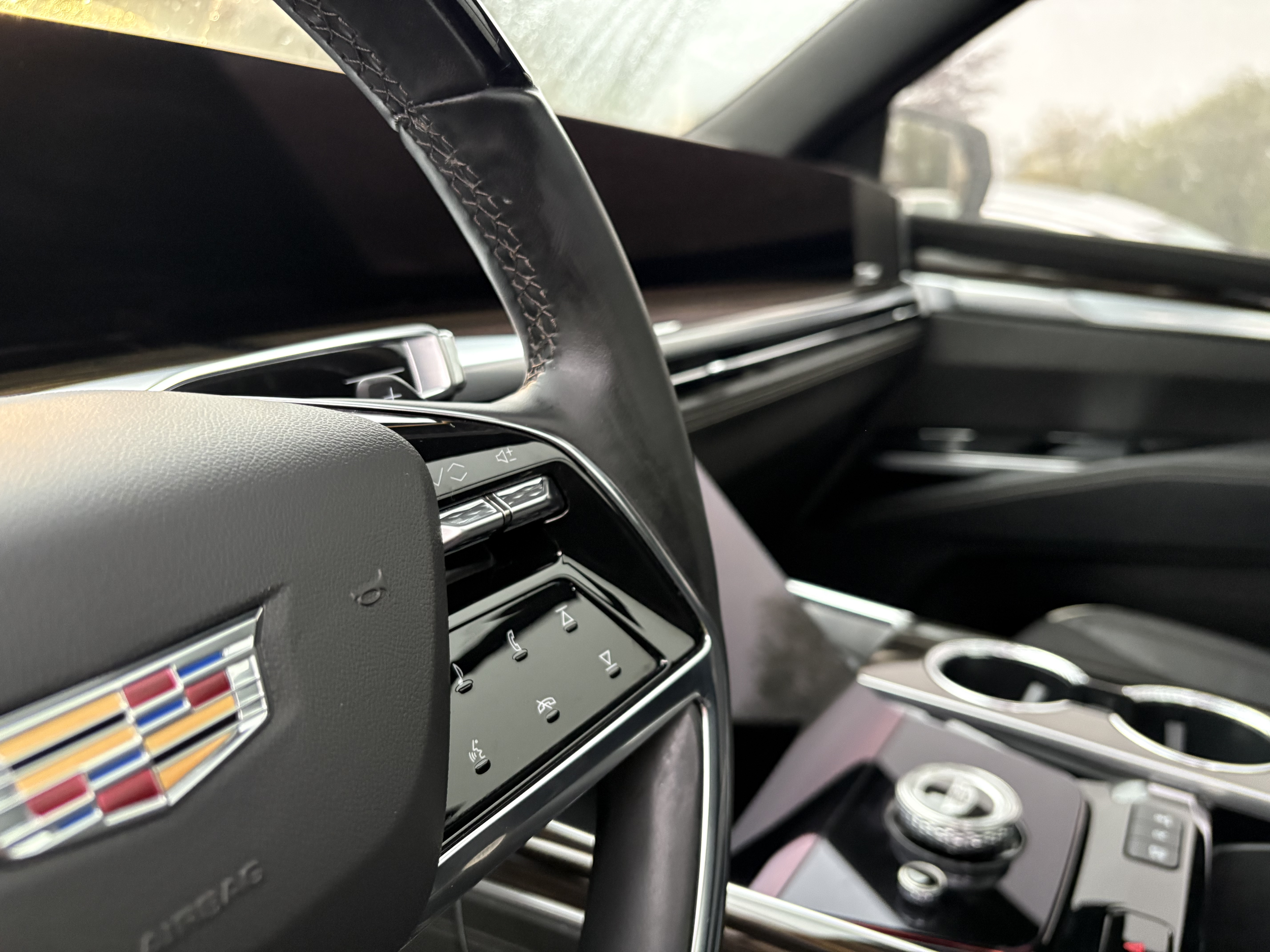 The interior of a car dashboard with various control buttons, an air vent, and a steering wheel partially visible, with a blurry view outside the windshield.