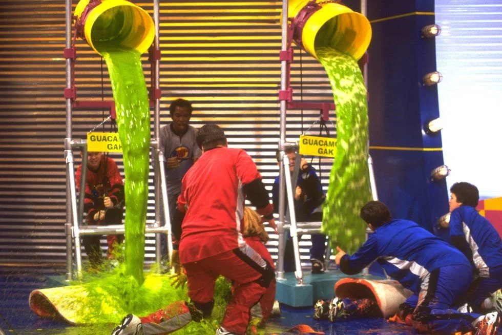 People in colourful athletic clothes are participating in a game show event with green slime flowing from two large yellow pipes.