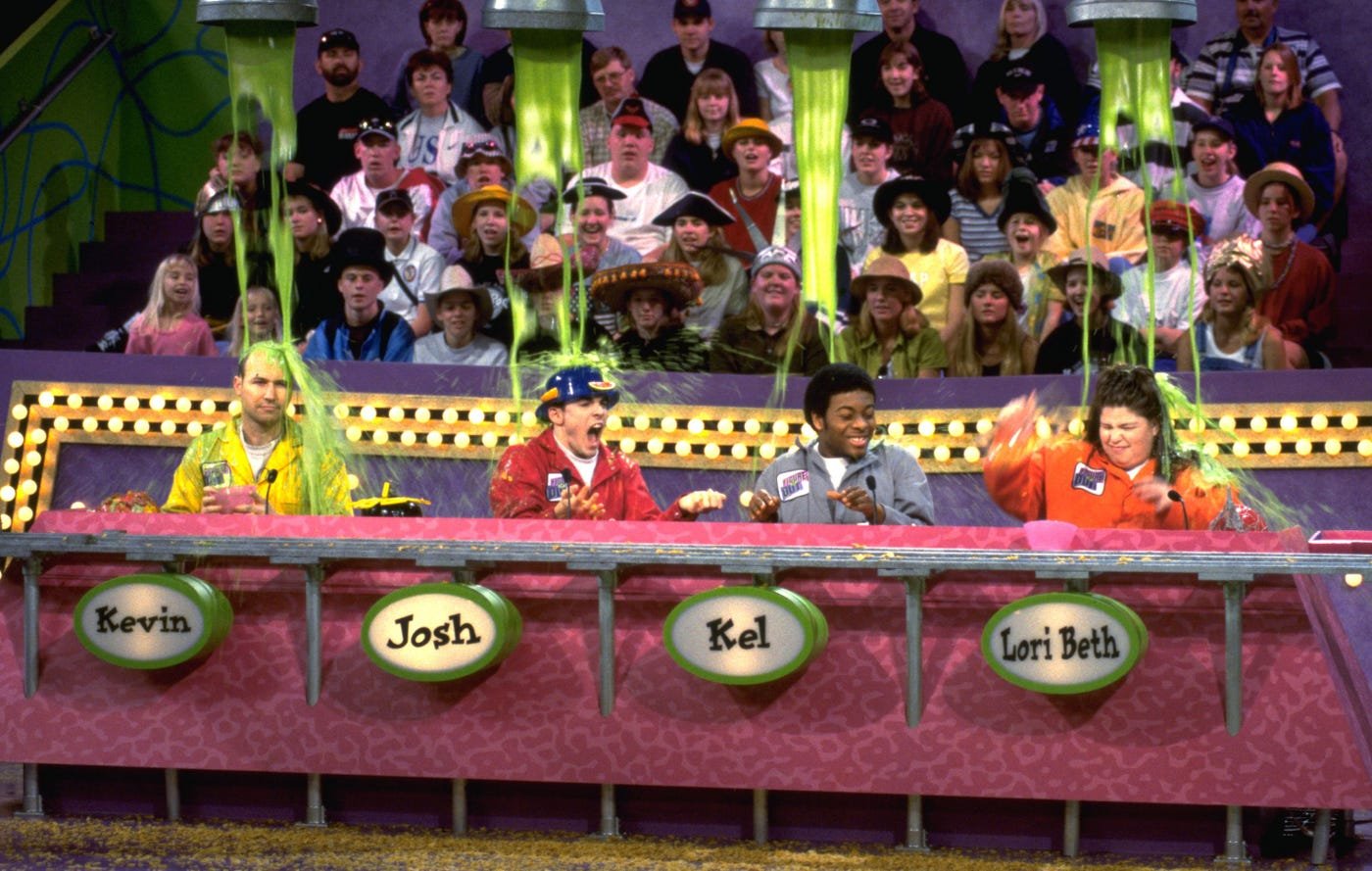 Four game show contestants sitting at a table with four green nameplates labeled Kevin, Josh, Kel, and Lori Beth, as green slime falls onto them. Audience members watch in the background.