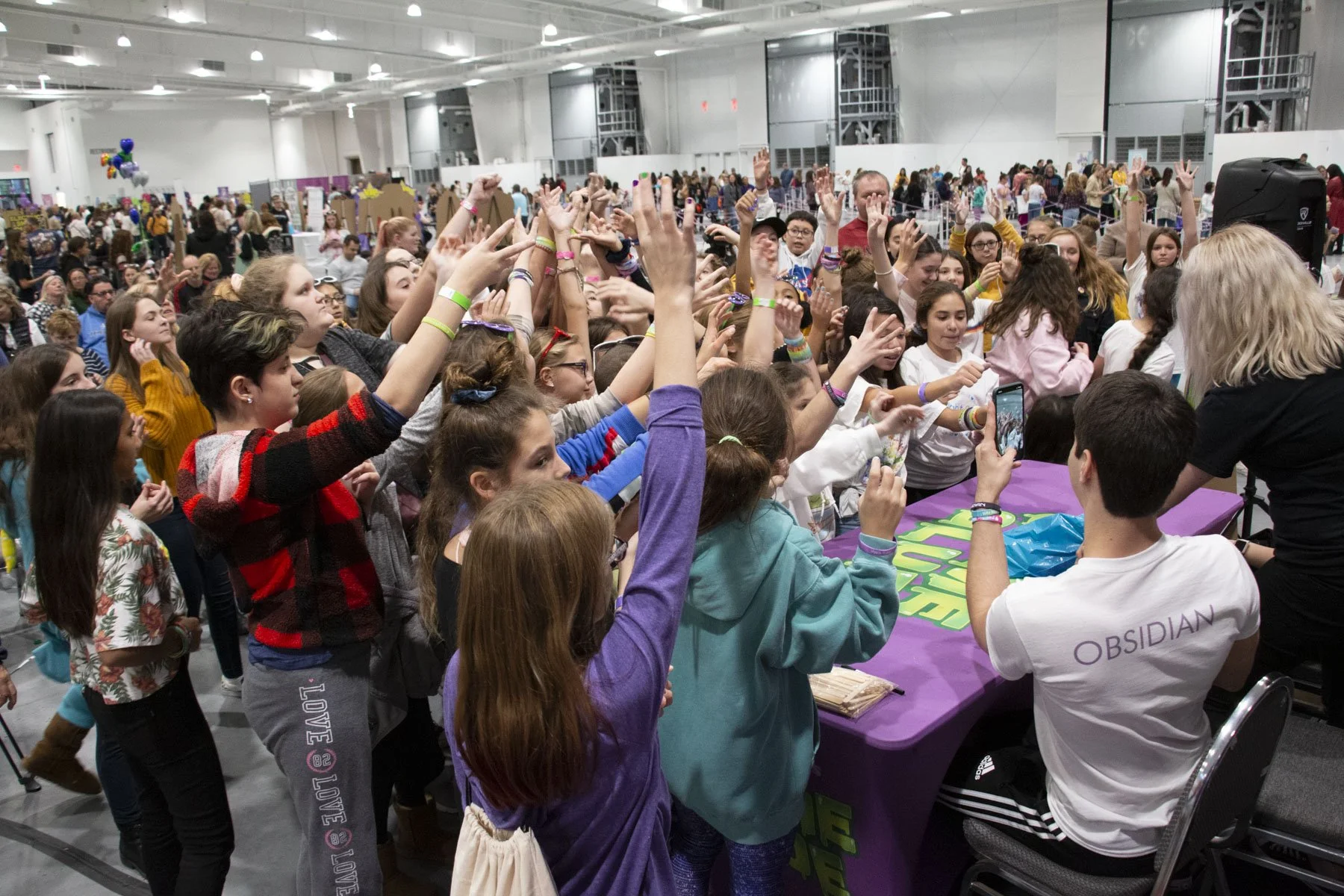 A large group of children and teenagers gathered at an indoor slime event, many with their hands raised, some taking photos. The scene has many booths and displays in the background, with a wide-open space and high ceilings.