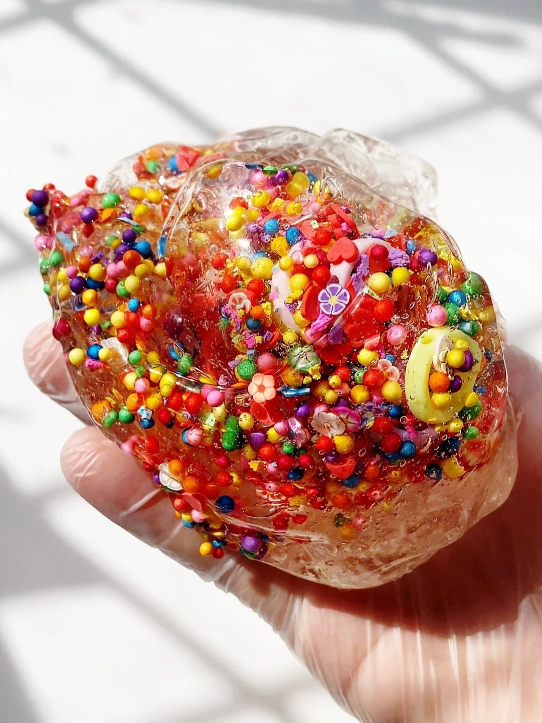 A hand wearing a glove holding a clear slime ball filled with colorful sprinkles and small decorative objects.