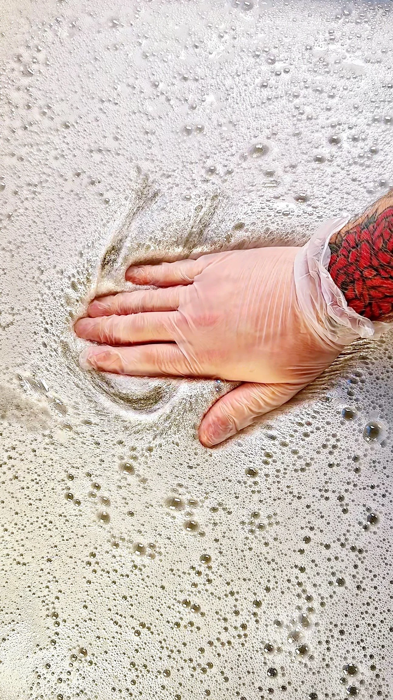A person's hand with a tattoo, wearing a latex glove, playing with a large batch of clear slime bubbles.