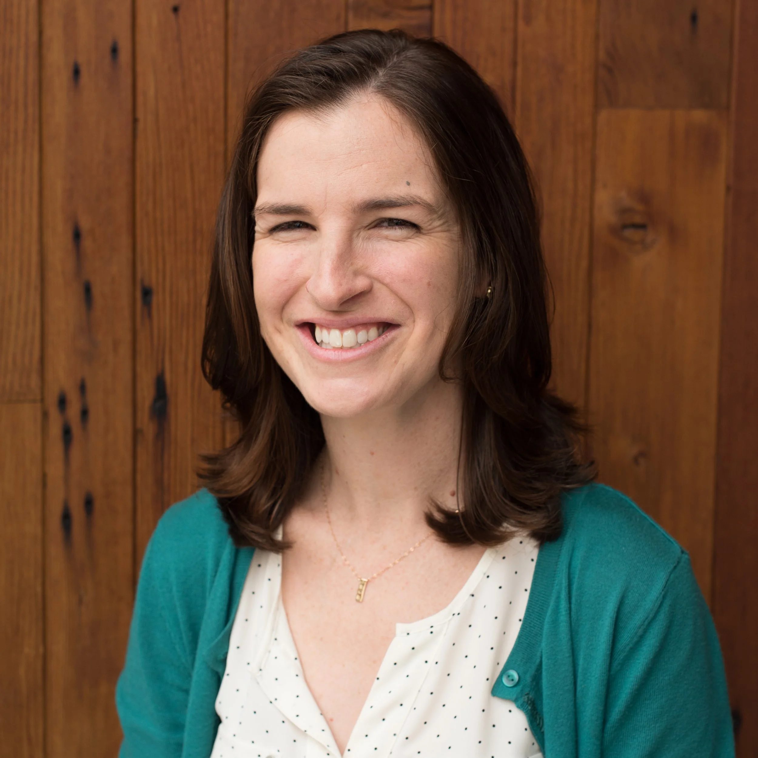 A woman with brown hair smiling, wearing a white polka dot blouse and a teal cardigan, standing in front of a wooden wall.