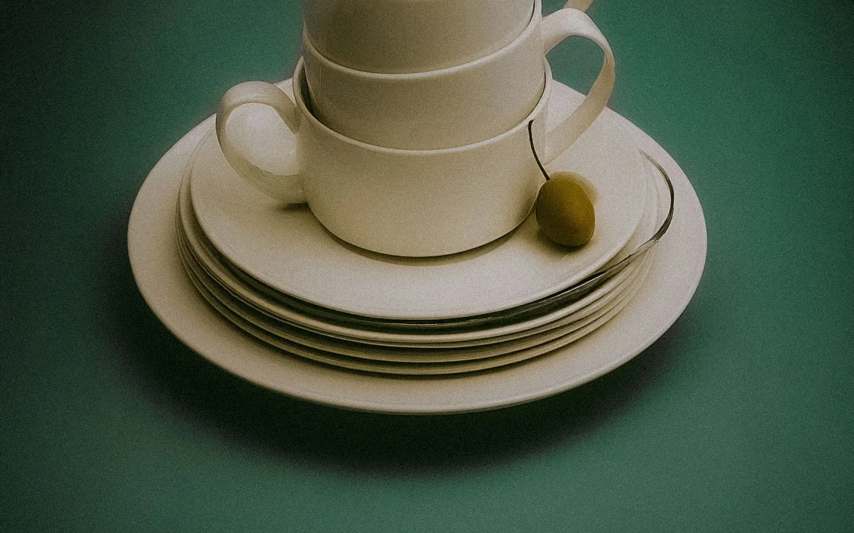 A stack of white porcelain cups and saucers on a white round plate, with a single green cherry resting on the edge of the plate, placed on a green table.