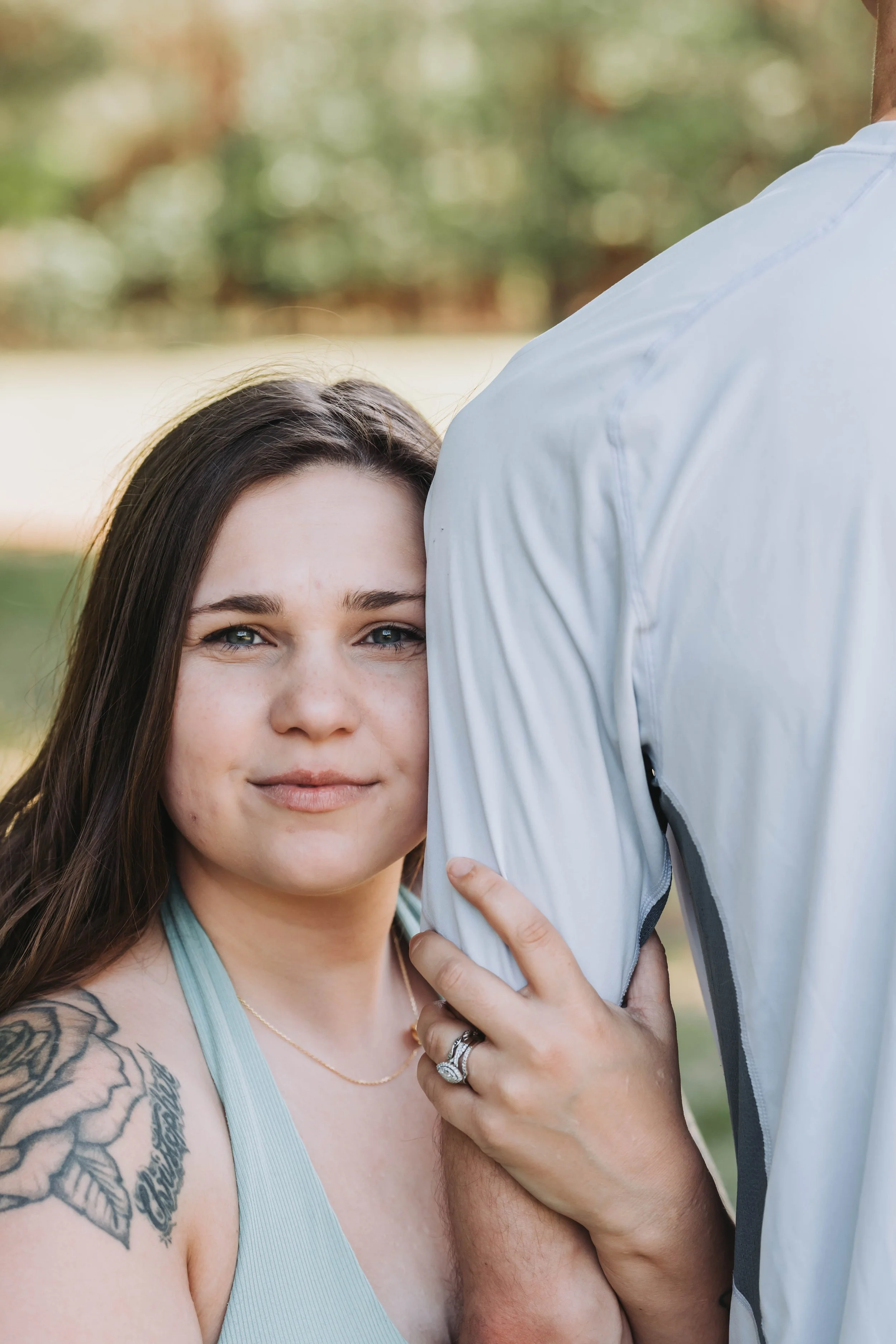 Close-up of a young woman with long brown hair, looking at the camera, with tattoos on her arm and wearing a light blue tank top. She is holding hands with a person wearing a white shirt, whose arm and hand are visible in the image. The woman has a gentle expression and is outdoors with blurred green and brown background.