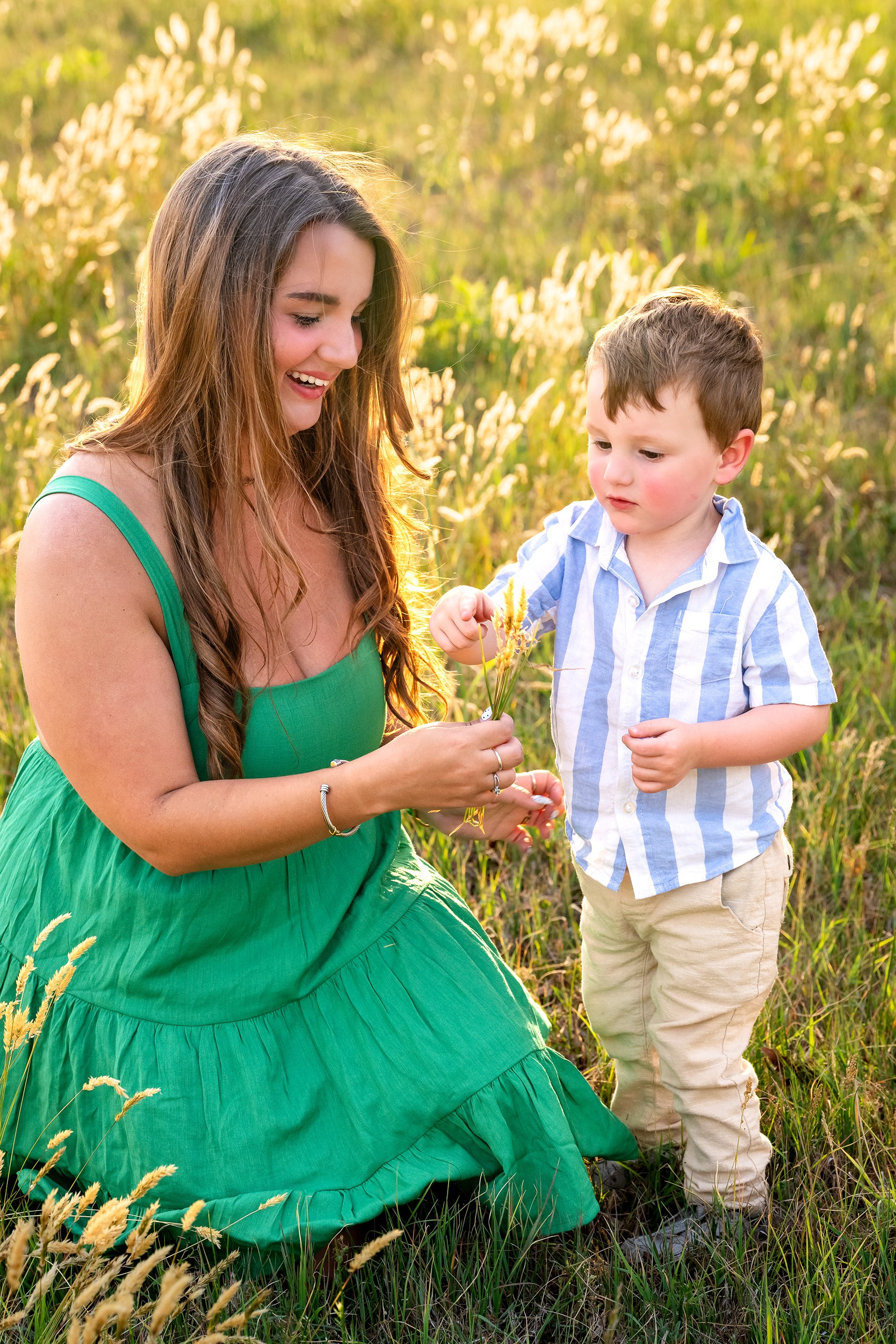 A woman in a green dress sitting in a field of tall grass and wildflowers, smiling and talking to a young boy in a blue and white striped shirt, who is holding a flower.