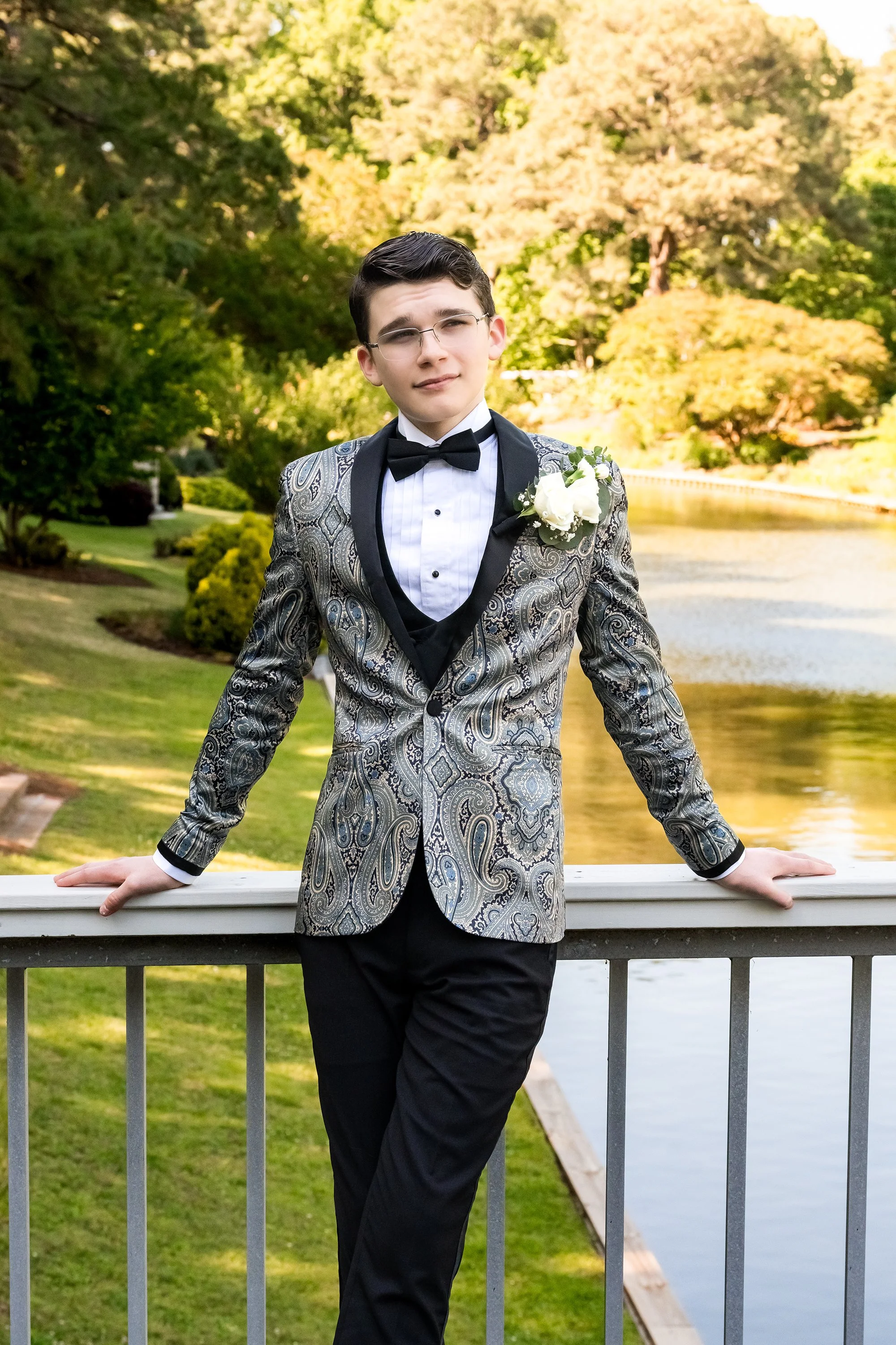 A young man in a patterned tuxedo jacket, white dress shirt, black bow tie, and boutonniere, standing outdoors by a river and lush foliage, posing with one hand on a bridge railing and the other relaxed, smiling gently.