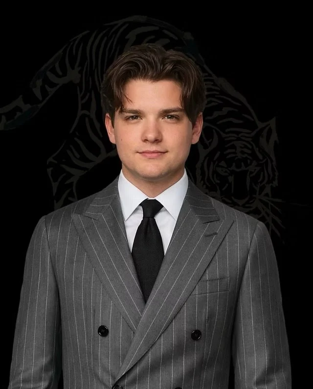 Young man in a gray pinstripe suit and black tie standing in front of a black background with a faint tiger illustration.