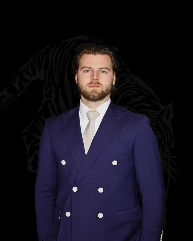 A man with brown hair, beard, and serious expression wearing a blue double-breasted suit with white buttons, white shirt, and white tie against a black background with a faint outline of a tiger.